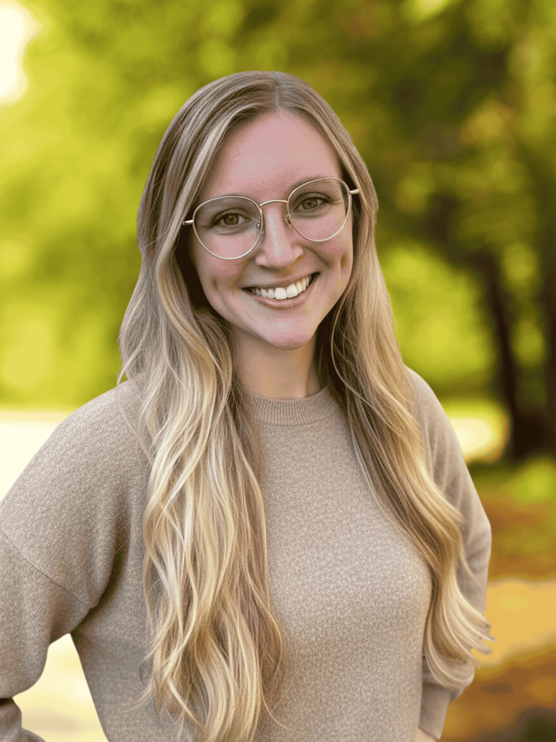 A young woman with long blonde hair wearing glasses and a beige sweater outdoors with green and yellow blurred trees in the background.