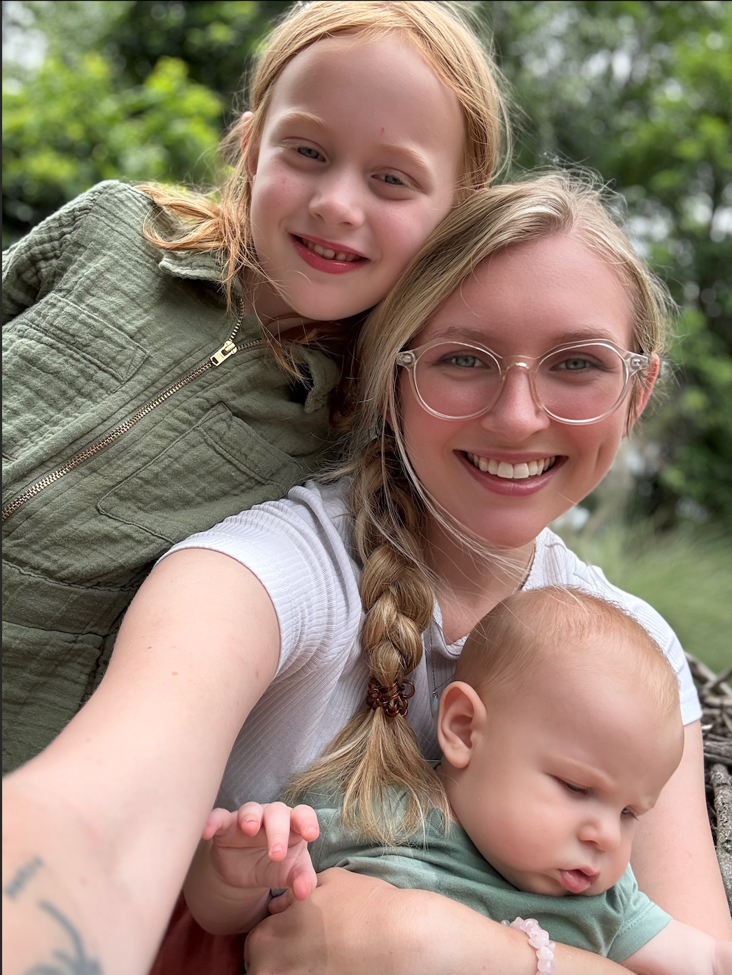 A woman with glasses and braided hair taking a selfie with two young children outdoors. The girl has red hair, and the baby has light hair, all smiling.