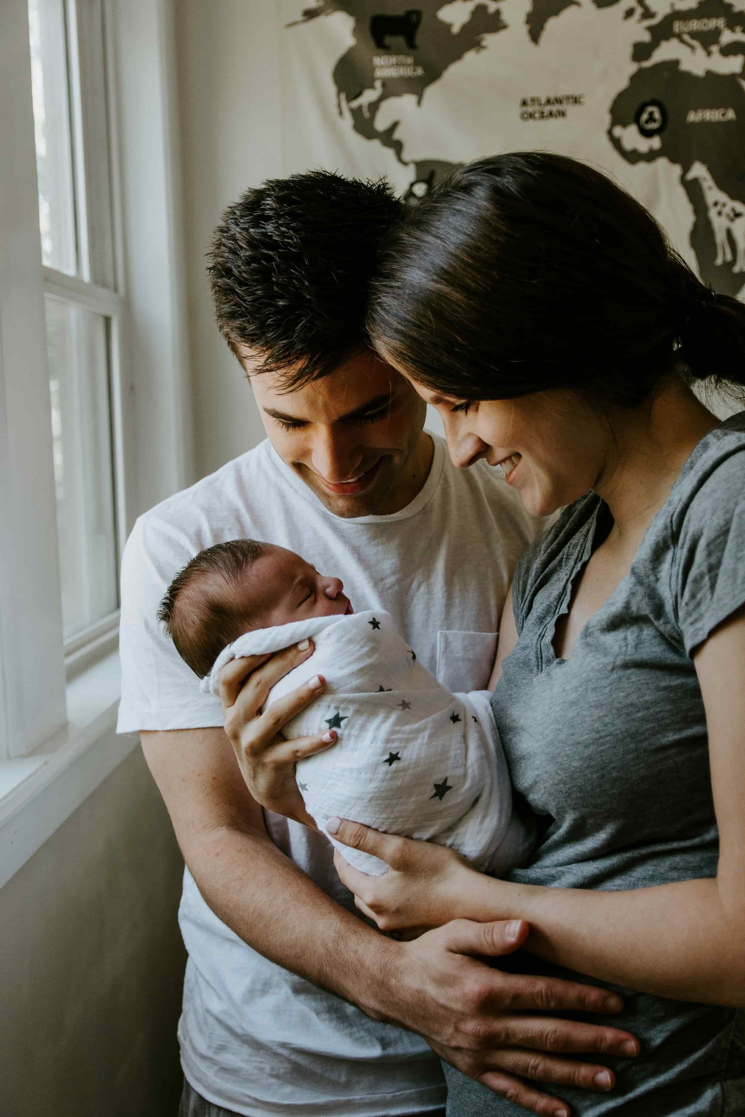 A couple holding a newborn baby near a window, smiling and looking at the baby, with a world map poster on the wall behind them.