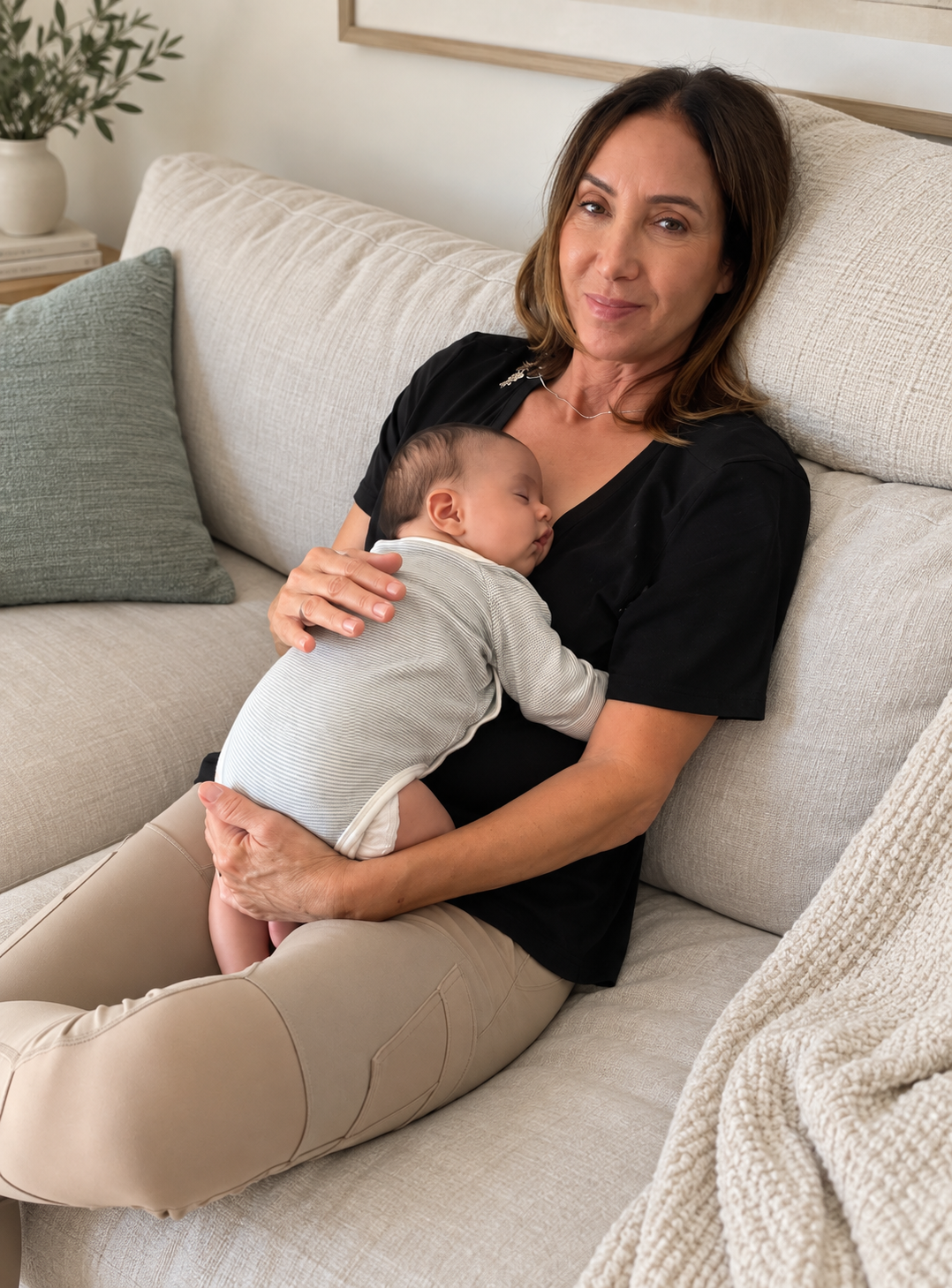 A woman with brown hair and a black shirt holding a sleeping baby on a beige sofa in a cozy living room.