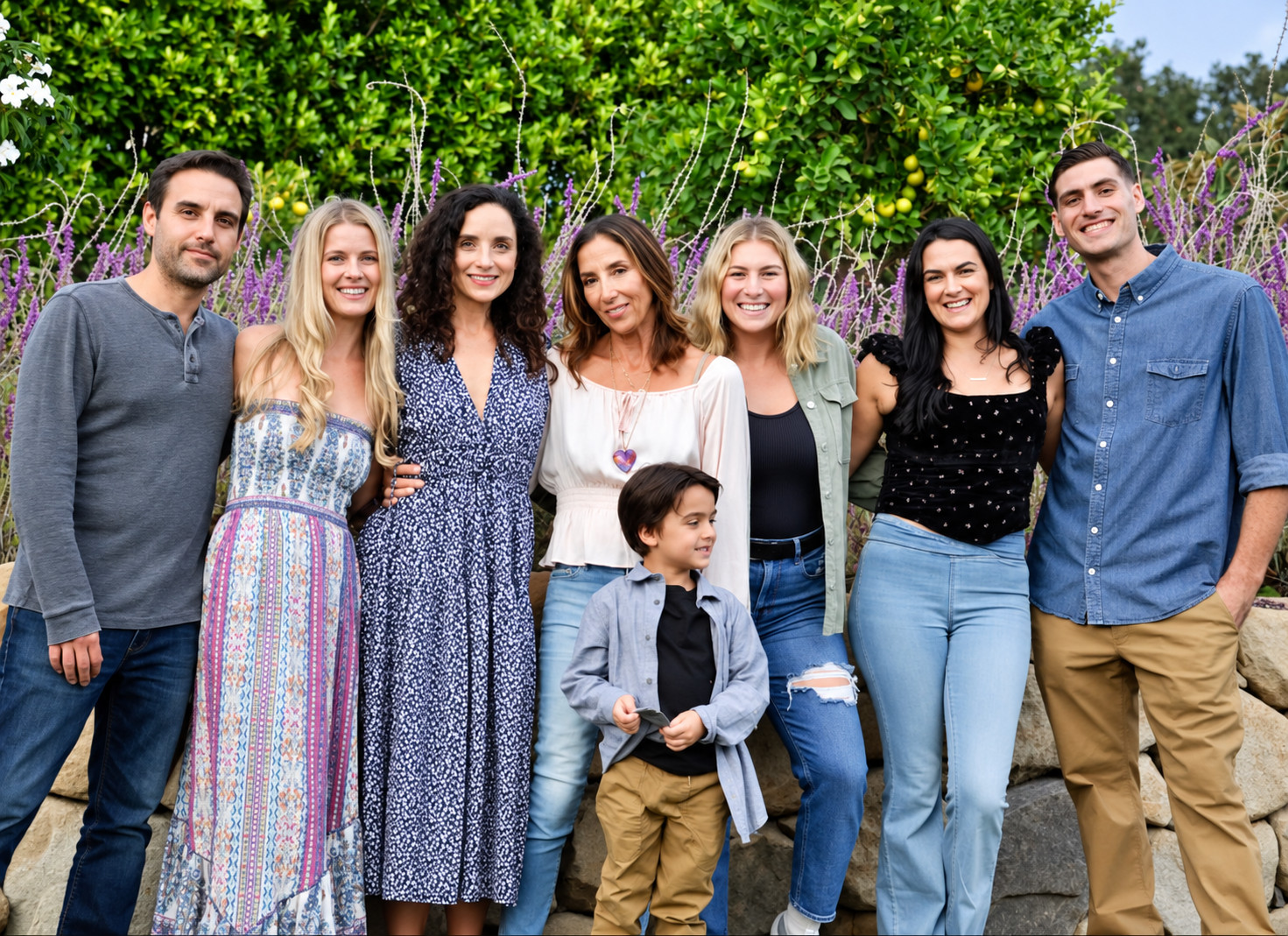 A diverse group of nine people, seven women, one man, and a young boy, standing outdoors in front of lush green bushes and purple flowering plants, smiling at the camera.