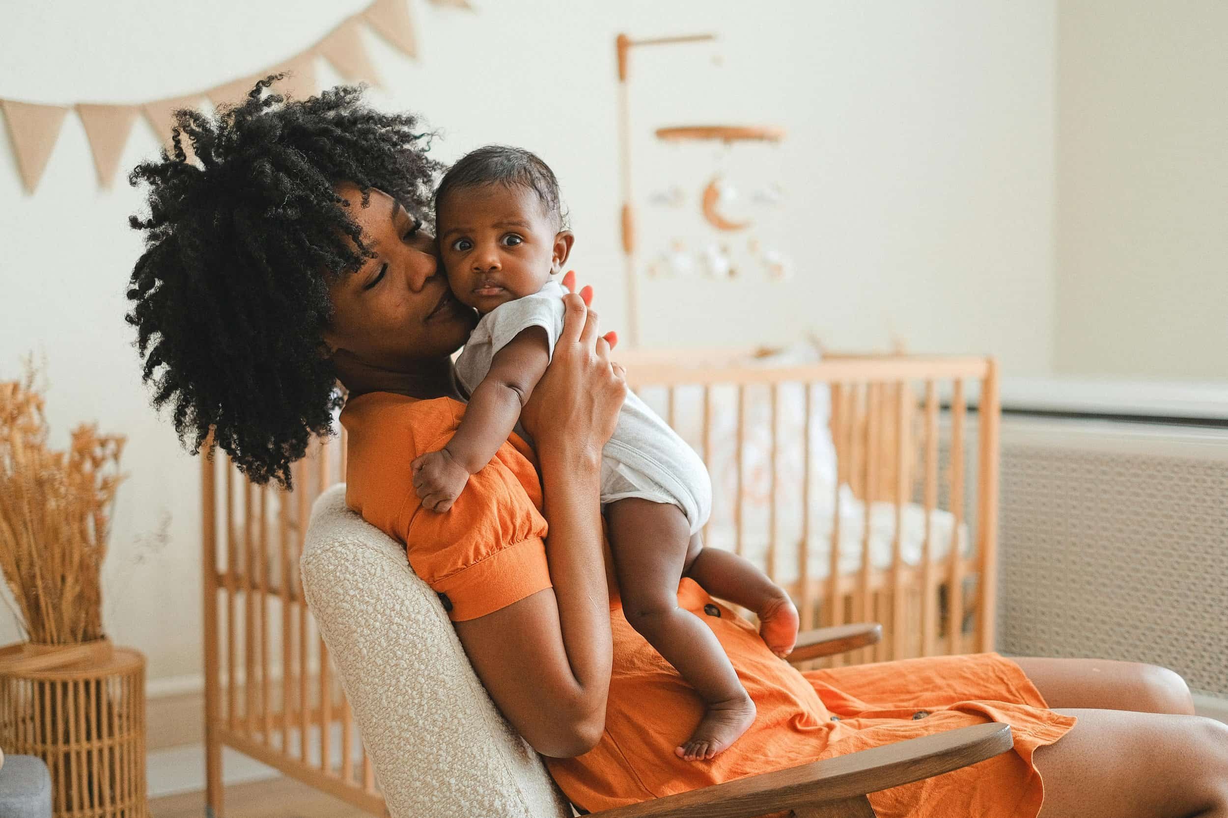 A woman with curly black hair holding a surprised baby in a nursery with a crib and decorative wall hanging.