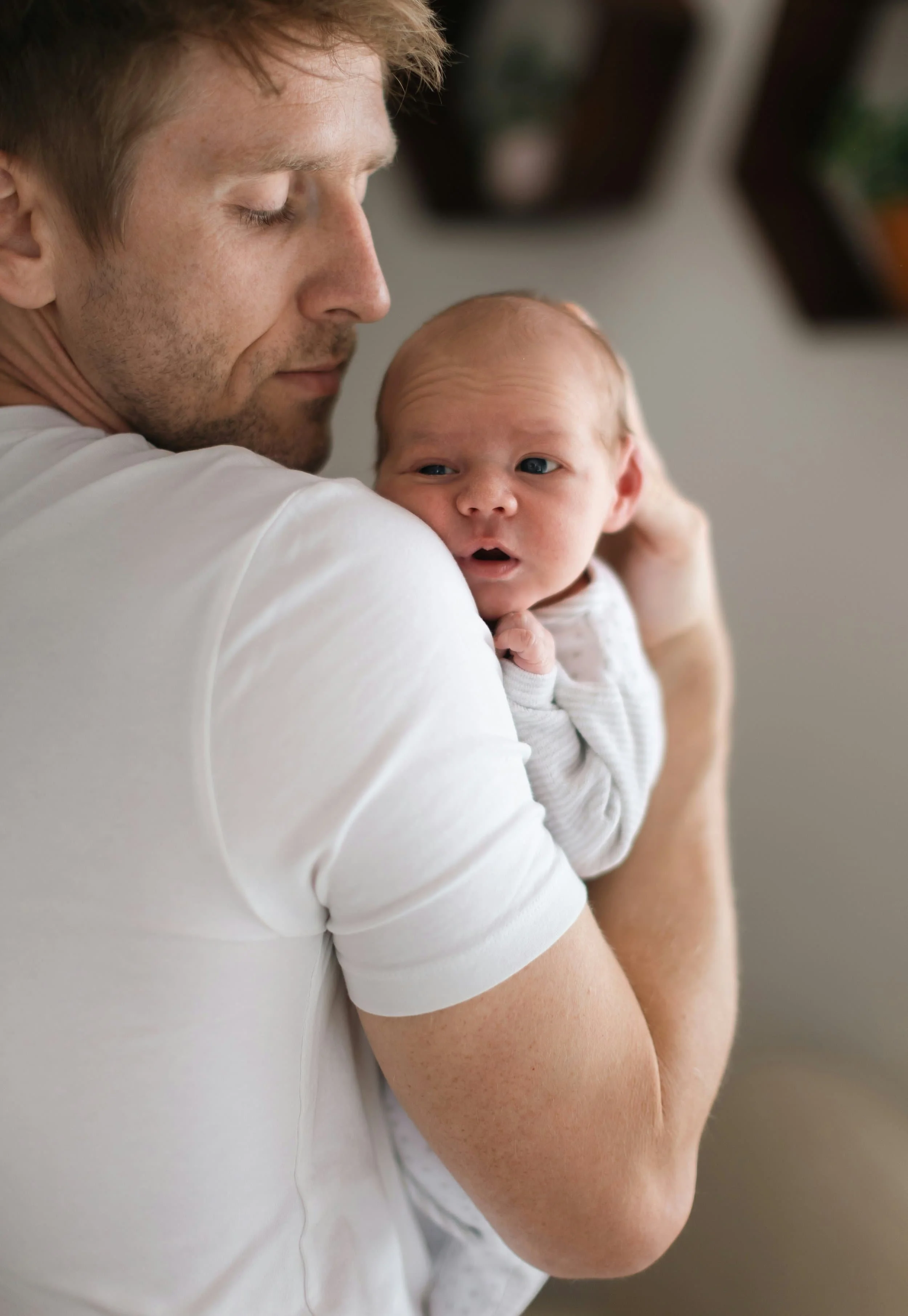 A man with red hair and light skin holding a newborn baby with red hair, blue eyes, and light skin, both in white clothing, with the man gently holding the baby close to his chest.