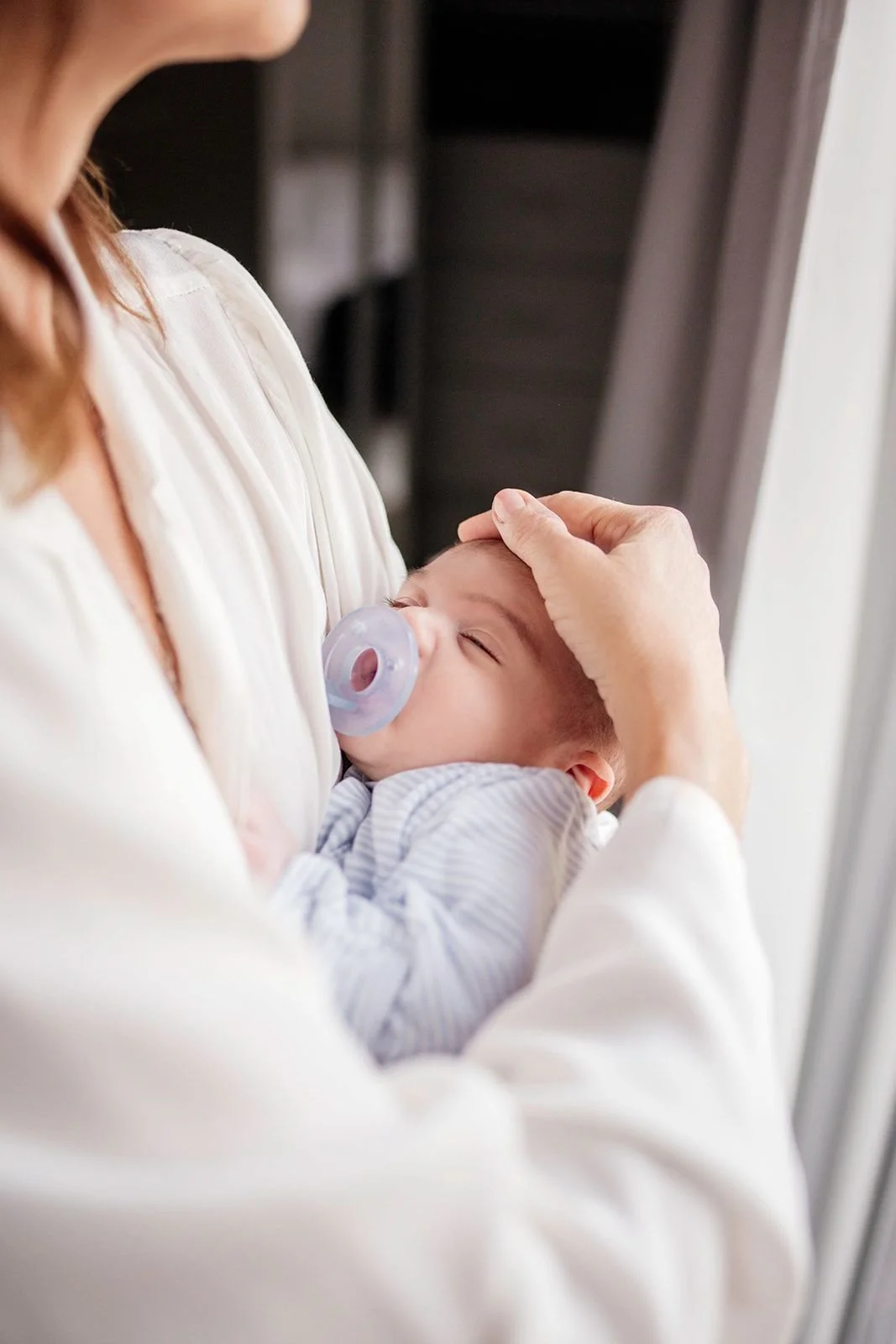 A woman cradles a sleeping baby with a pacifier, gently patting the baby's head. The image captures a tender moment of care and comfort.