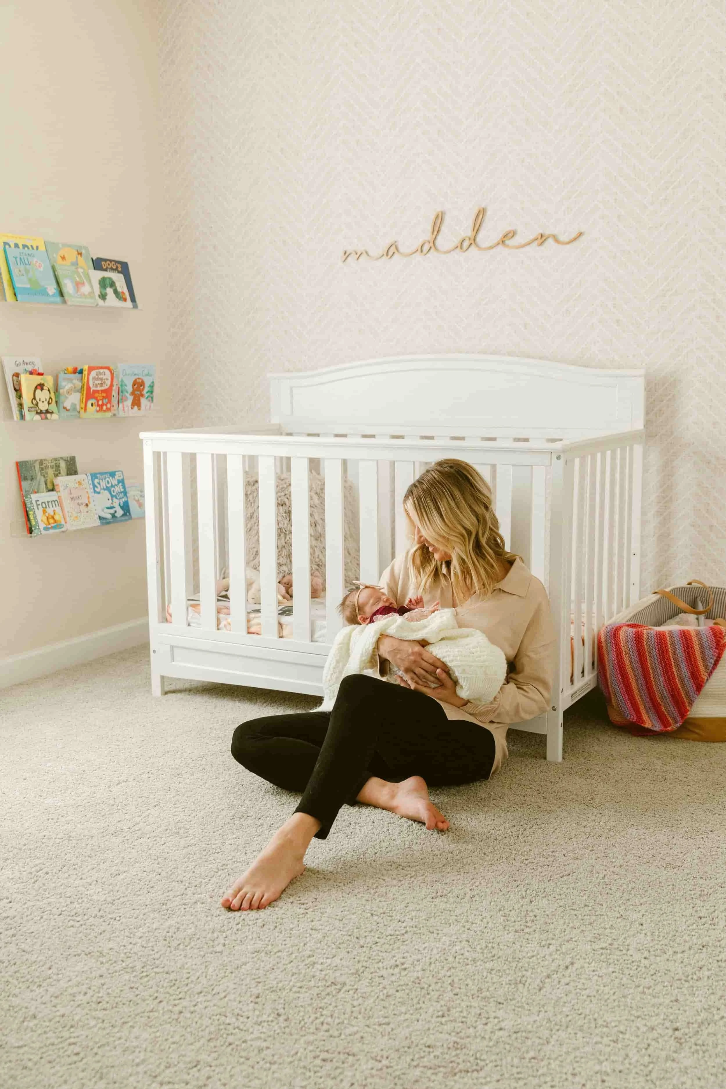 A woman sitting on the floor with crossed legs in front of a white crib, holding a newborn baby. The nursery has a beige carpet, a wall with a textured pattern, and a bookshelf with children's books. The word 'modern' is written on the wall in cursive.