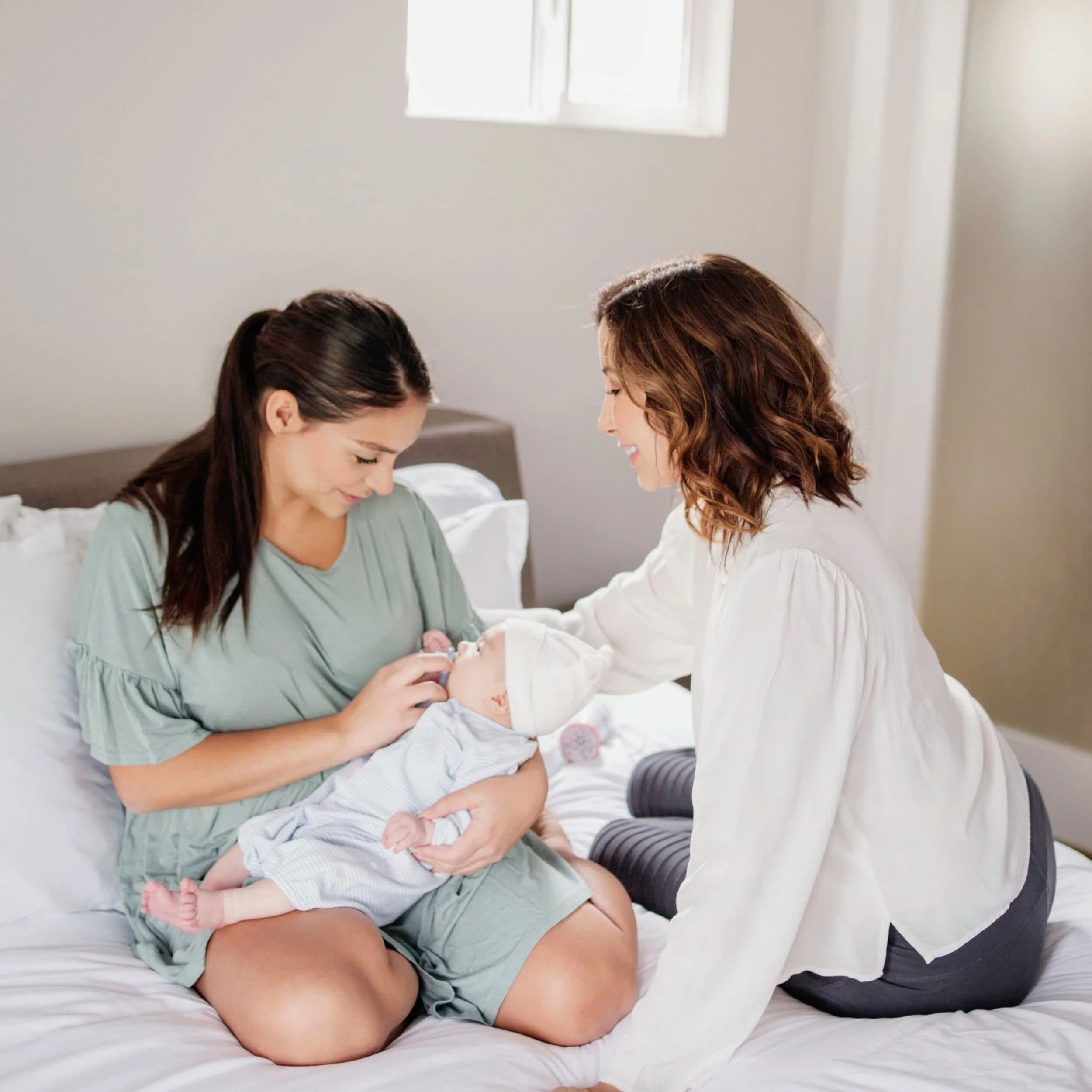Two women sitting on a bed in a hospital room, holding a newborn baby, smiling and looking lovingly at the infant.