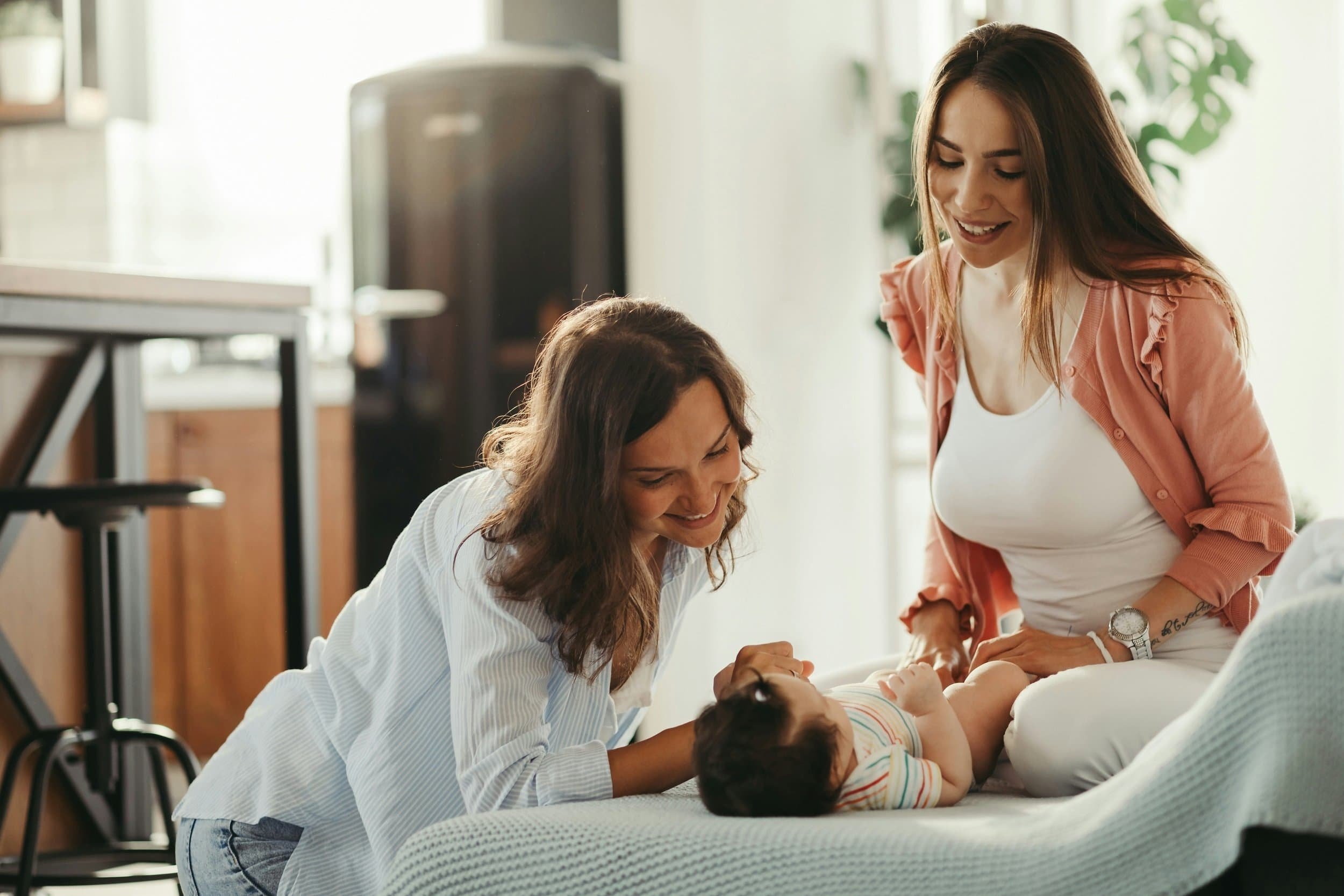 Two women are smiling and holding a young child's hands, playing on a bed in a bright room with sunlight coming through the windows.