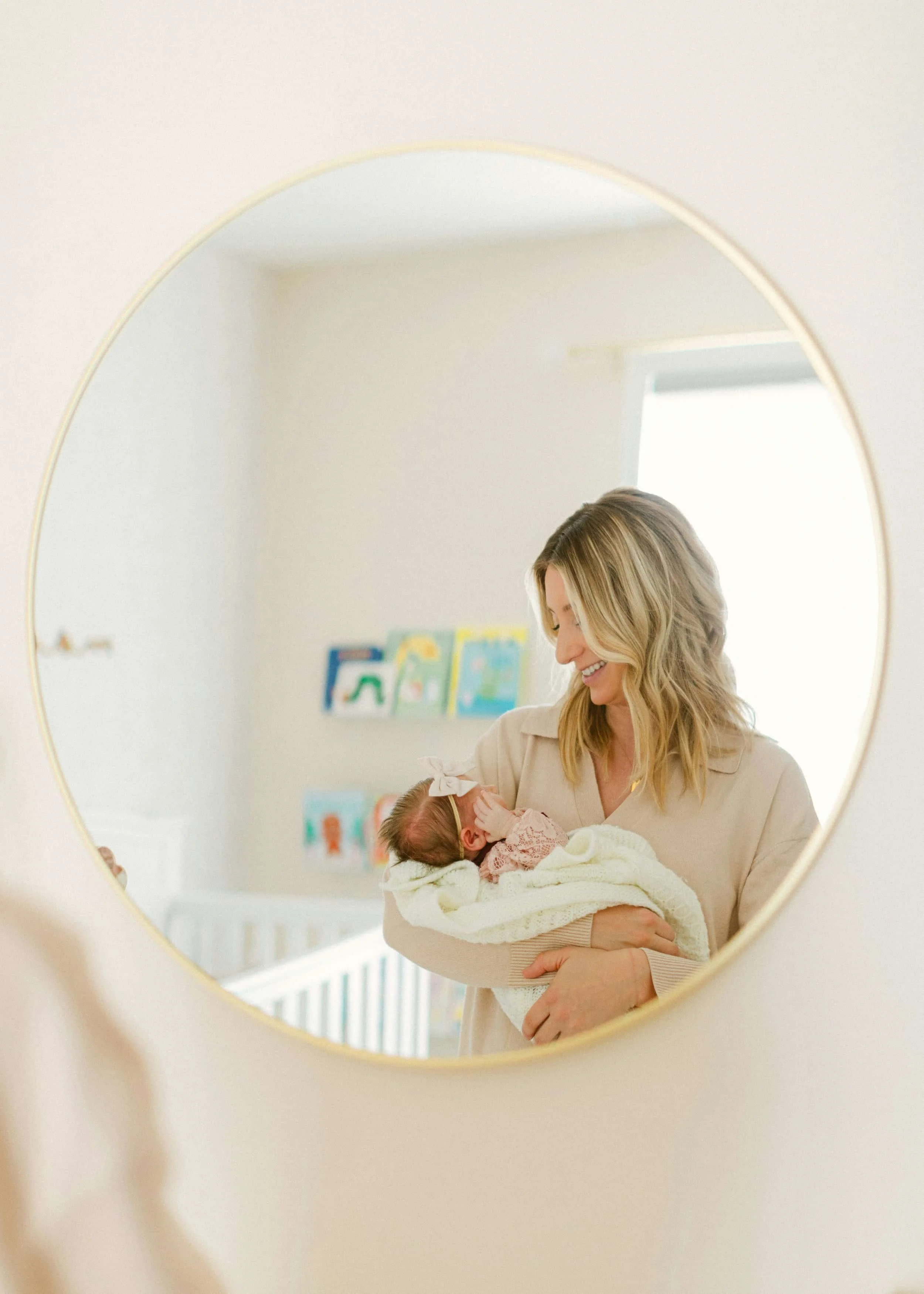 A woman holding a baby, seen through a circular mirror, smiling and looking at the baby, in a brightly lit room with framed artwork on the wall.