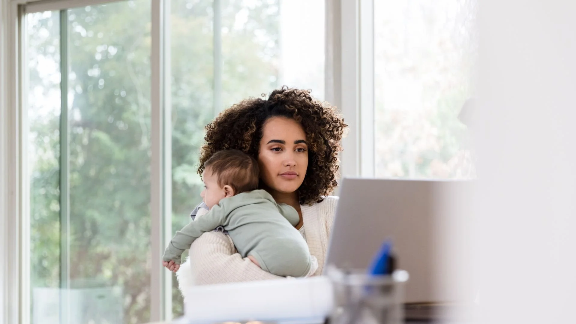 Mother holding sleeping baby while on laptop, representing Om Baby's virtual postpartum doula support in Los Angeles