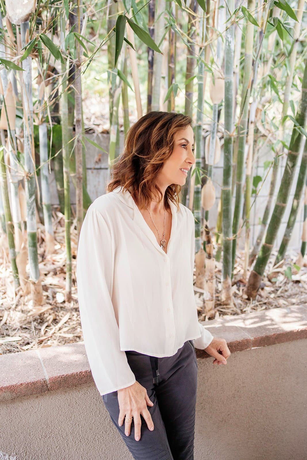 A woman with shoulder-length wavy brown hair standing outdoors near bamboo plants, wearing a cream blouse and dark pants.