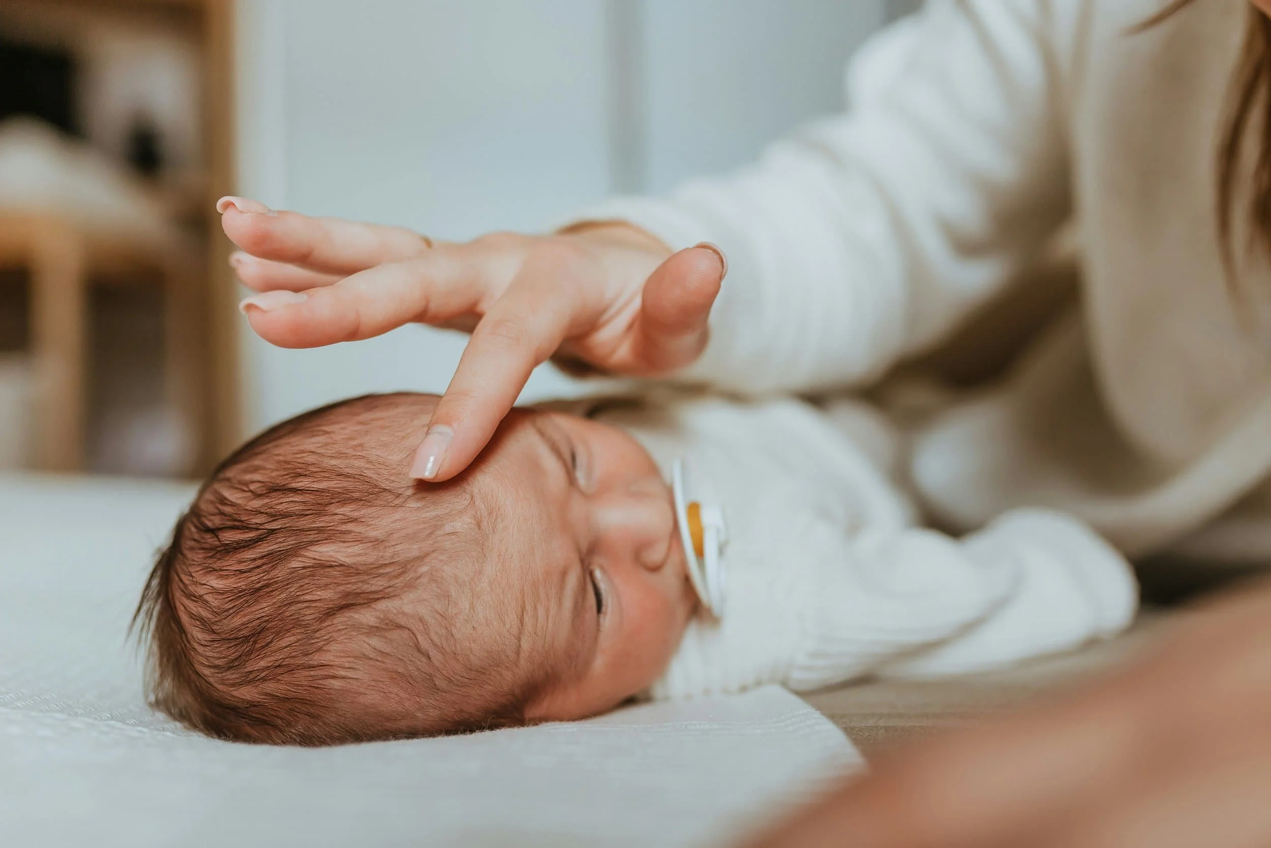 A newborn baby with dark hair lying on a soft surface and wearing a nasal oxygen tube. An adult's hand is gently touching the baby's head with their index finger.