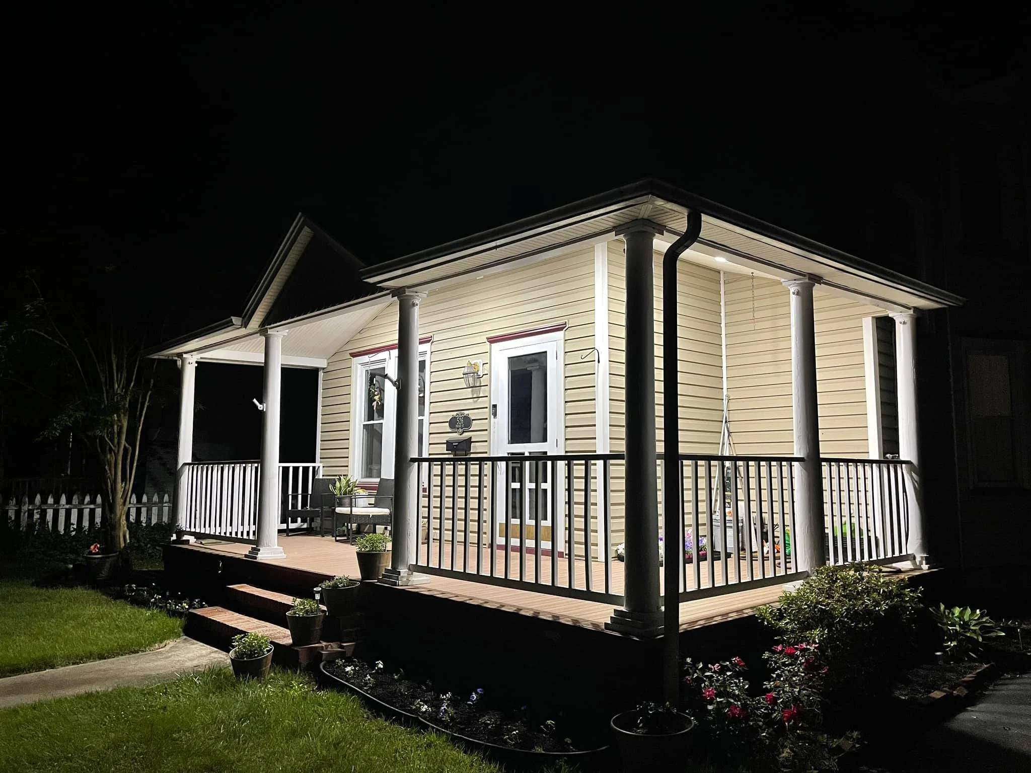 View of a well-lit porch and house at night, with yellow siding, white columns, railing, and potted plants.