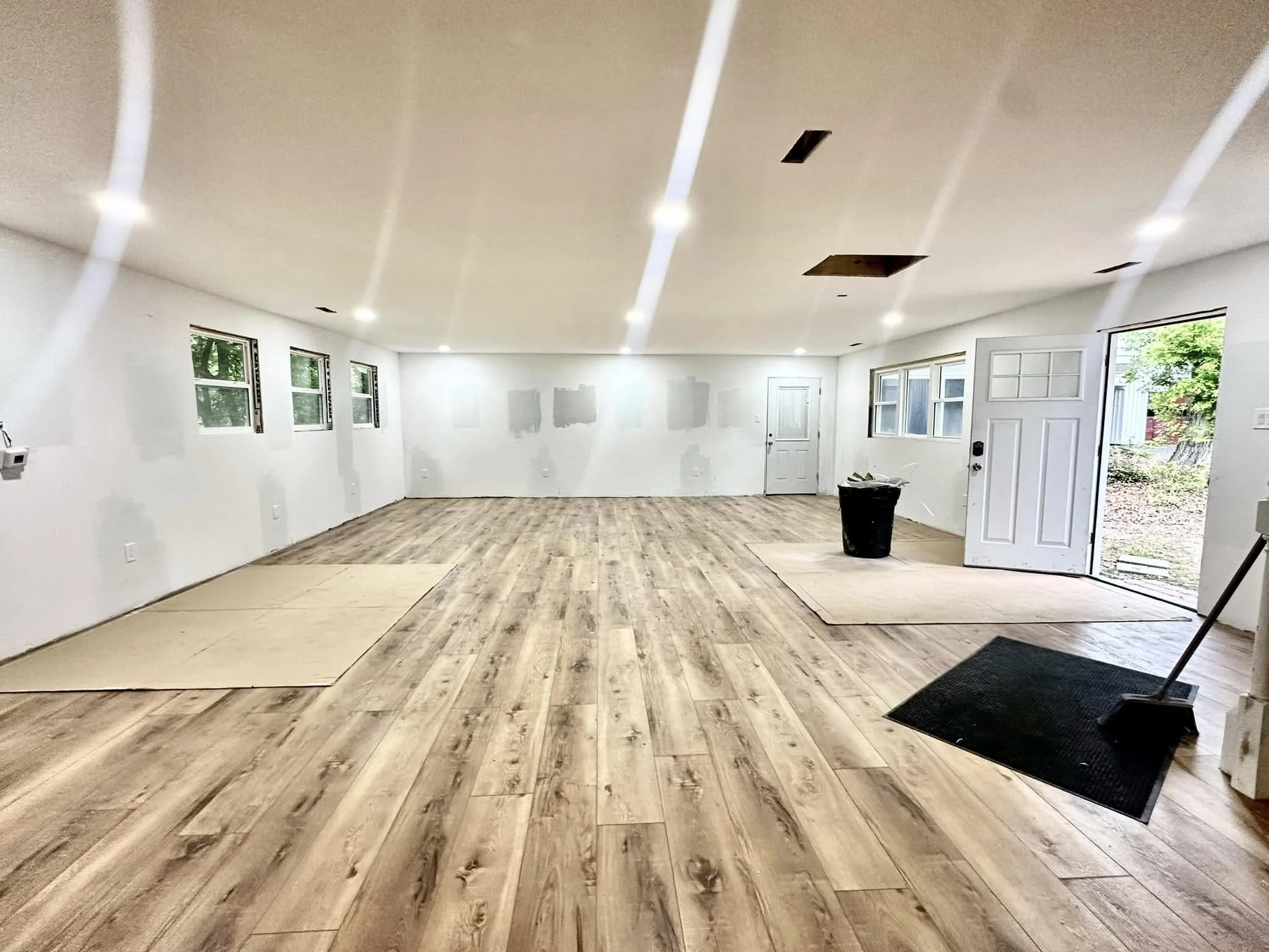 A spacious, unfinished room with white walls, wood flooring, multiple small windows, and an open door leading outside. The ceiling has recessed lighting and some openings for fixtures or vents. There are construction materials and a trash can present