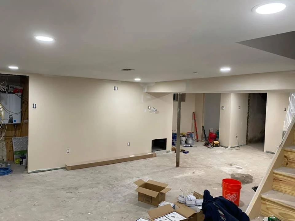 Interior of a basement under construction with drywall, ceiling lights, construction tools, and an unfinished floor.
