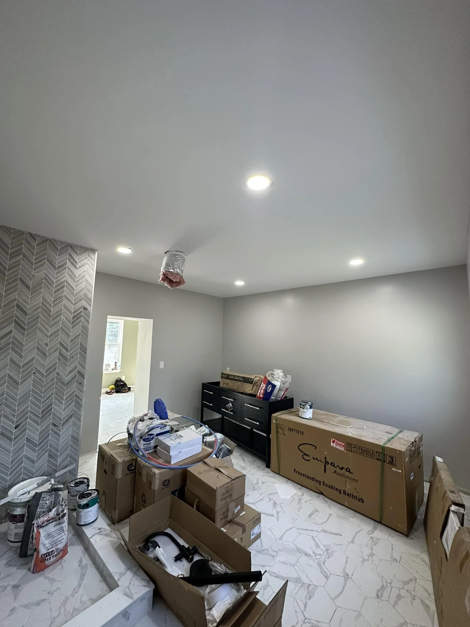 Room under renovation with boxes, tools, and a bathtub, with ceiling lights and a partially finished wall.