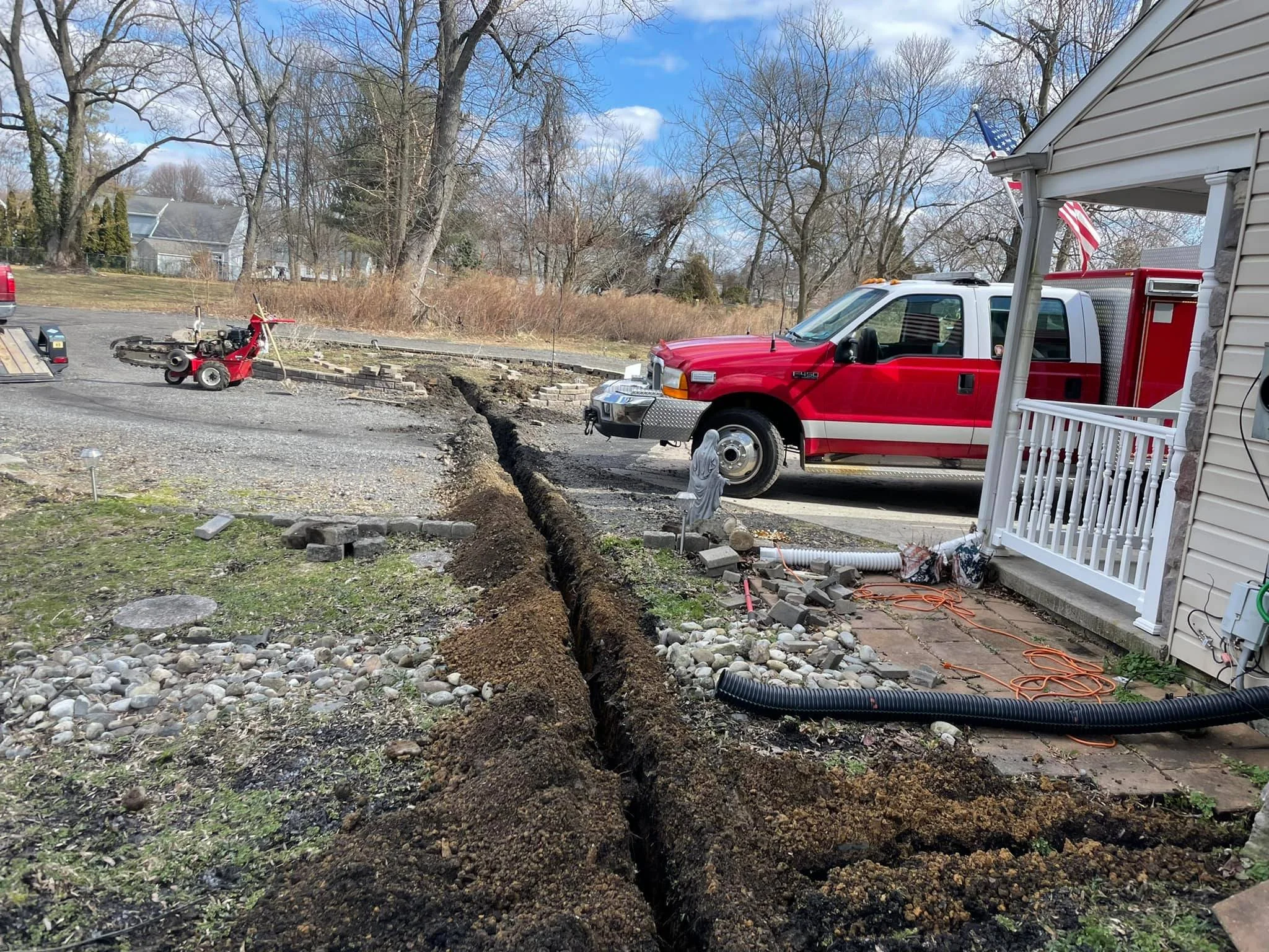 A freshly dug trench runs from the foreground toward the background next to a house with a porch. Construction equipment and tools are nearby, and in the background, there are leafless trees, a blue sky with clouds, and a parked red fire truck.