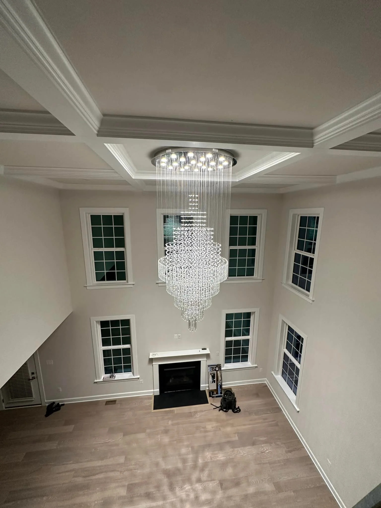 Interior view of a spacious living room with a large, elegant chandelier hanging from a tray ceiling, four tall windows with grid patterns, a fireplace with a black hearth, and hardwood floors.