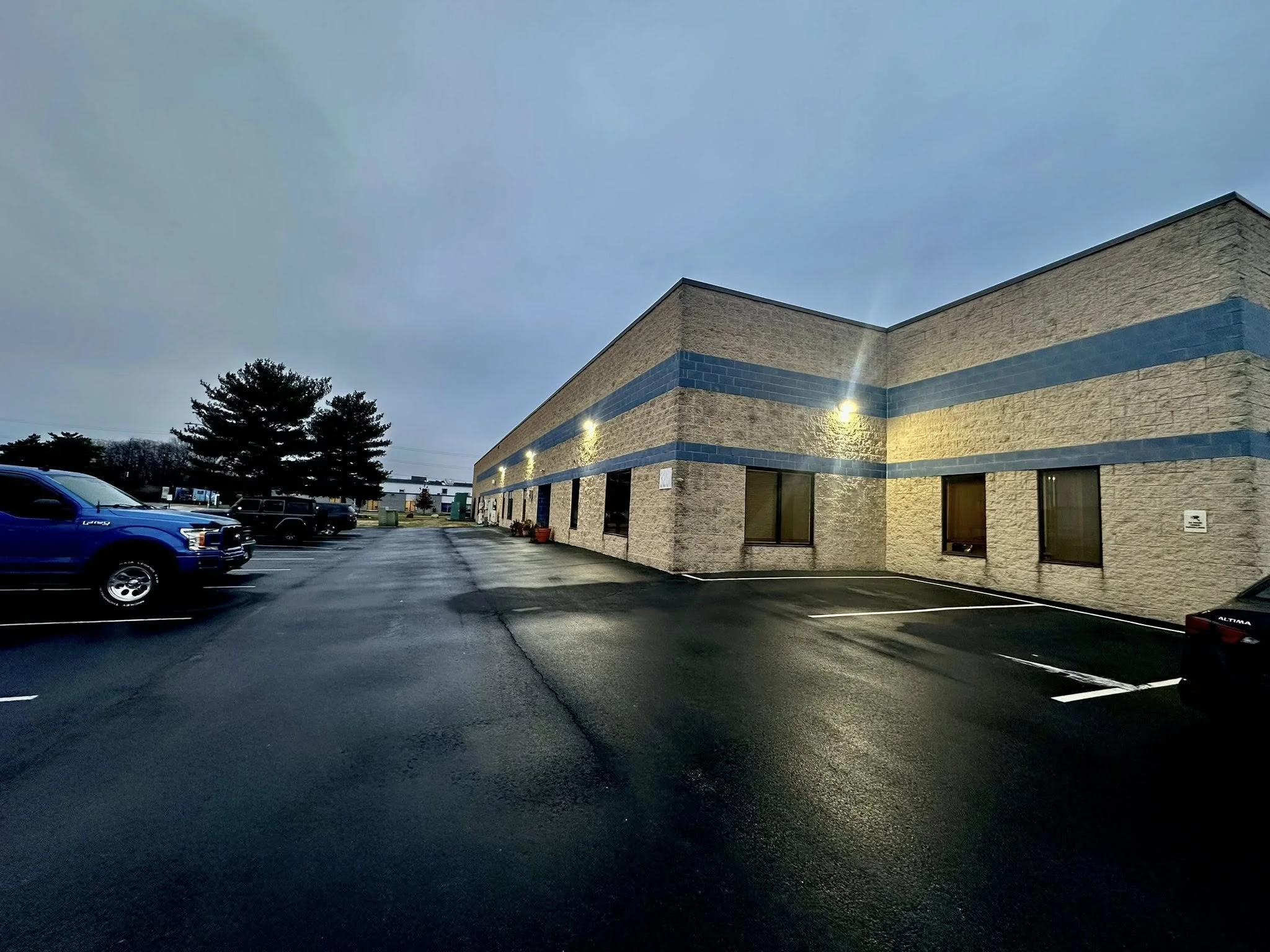 Parking lot outside a beige commercial building at dusk with several cars parked and lights illuminating the building.