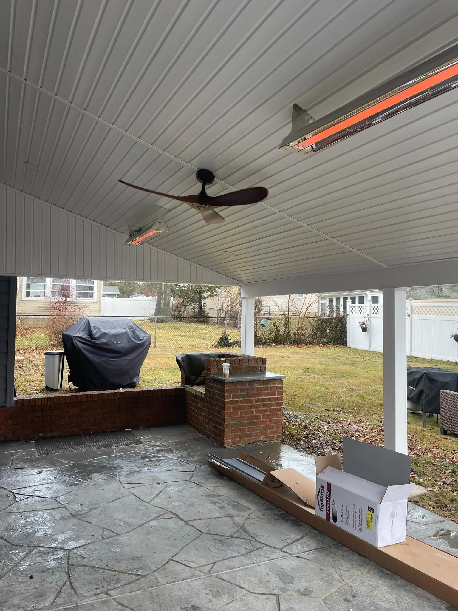 Covered outdoor patio with ceiling fan, two ceiling heaters, and a concrete floor. There is a brick ledge with a beverage on it, a cardboard box and some long items on the ground, with a fenced backyard in the background containing a covered grill an