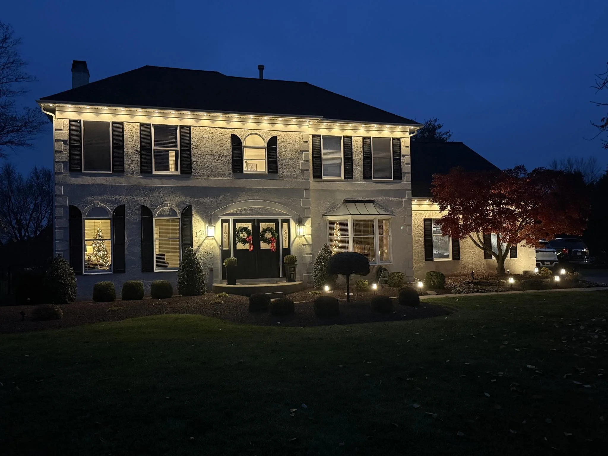 Two-story house decorated for Christmas with lights on the roofline, wreaths on the front door, and a decorated Christmas tree visible through the front window. The house has black shutters, a bay window, and a well-lit yard with bushes and a red-lea