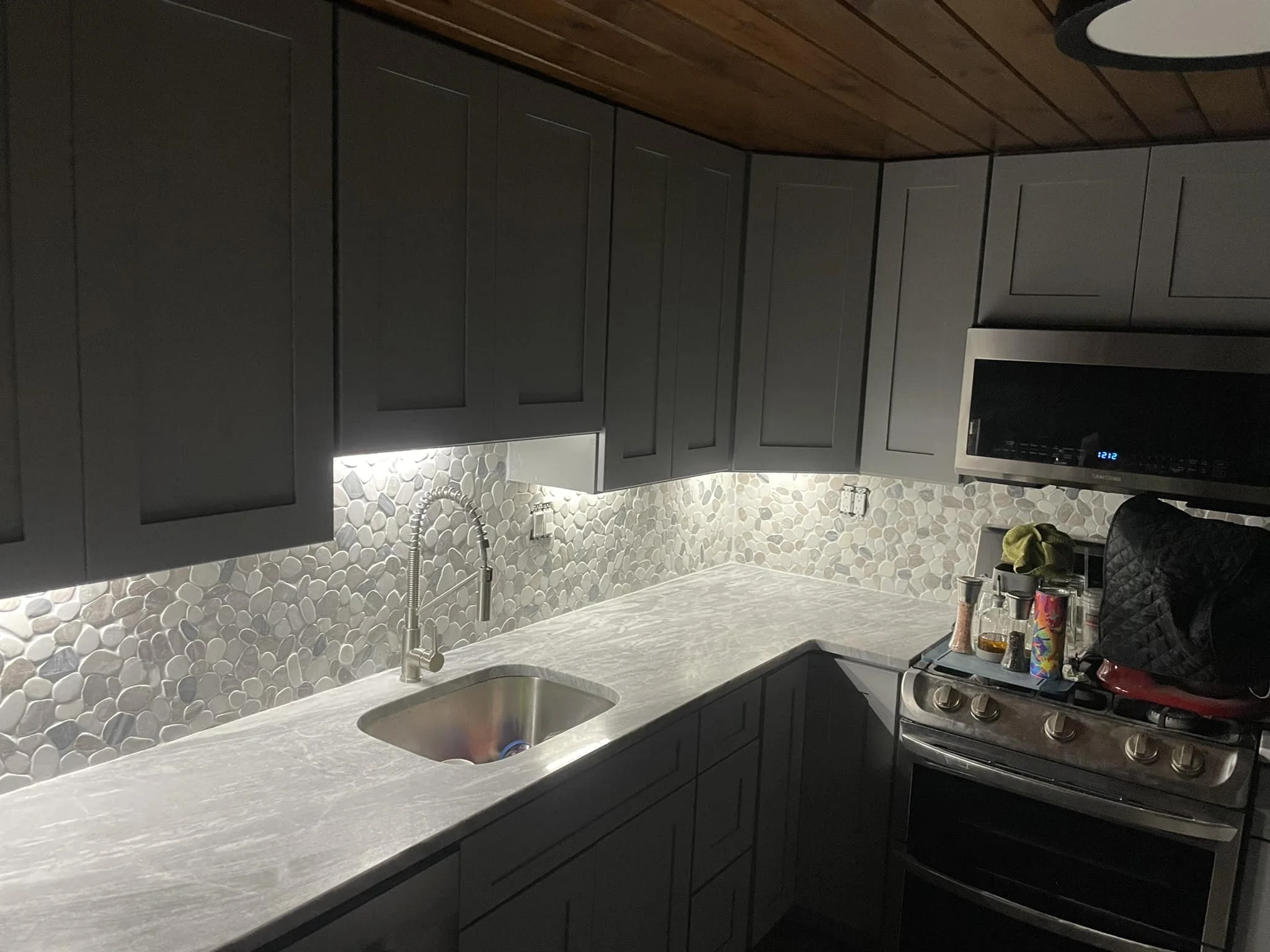 A modern kitchen with gray cabinets, a white marble countertop, a pebble backsplash, a stainless steel sink with a gooseneck faucet, a microwave oven, and a gas stove with some bottles and kitchen items on top.