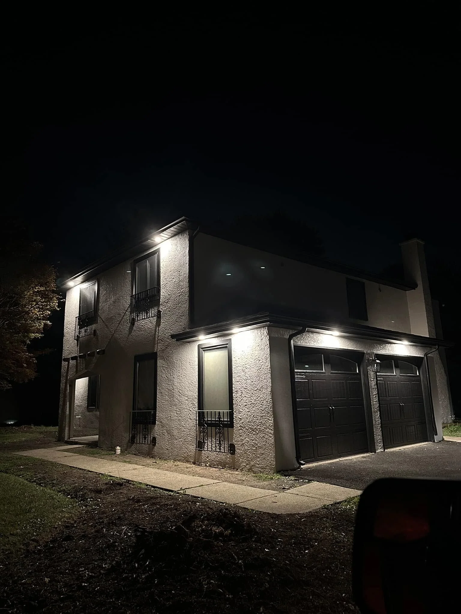 A two-story house with a garage, illuminated by exterior lights at night, with a sidewalk in front and a dark sky above.