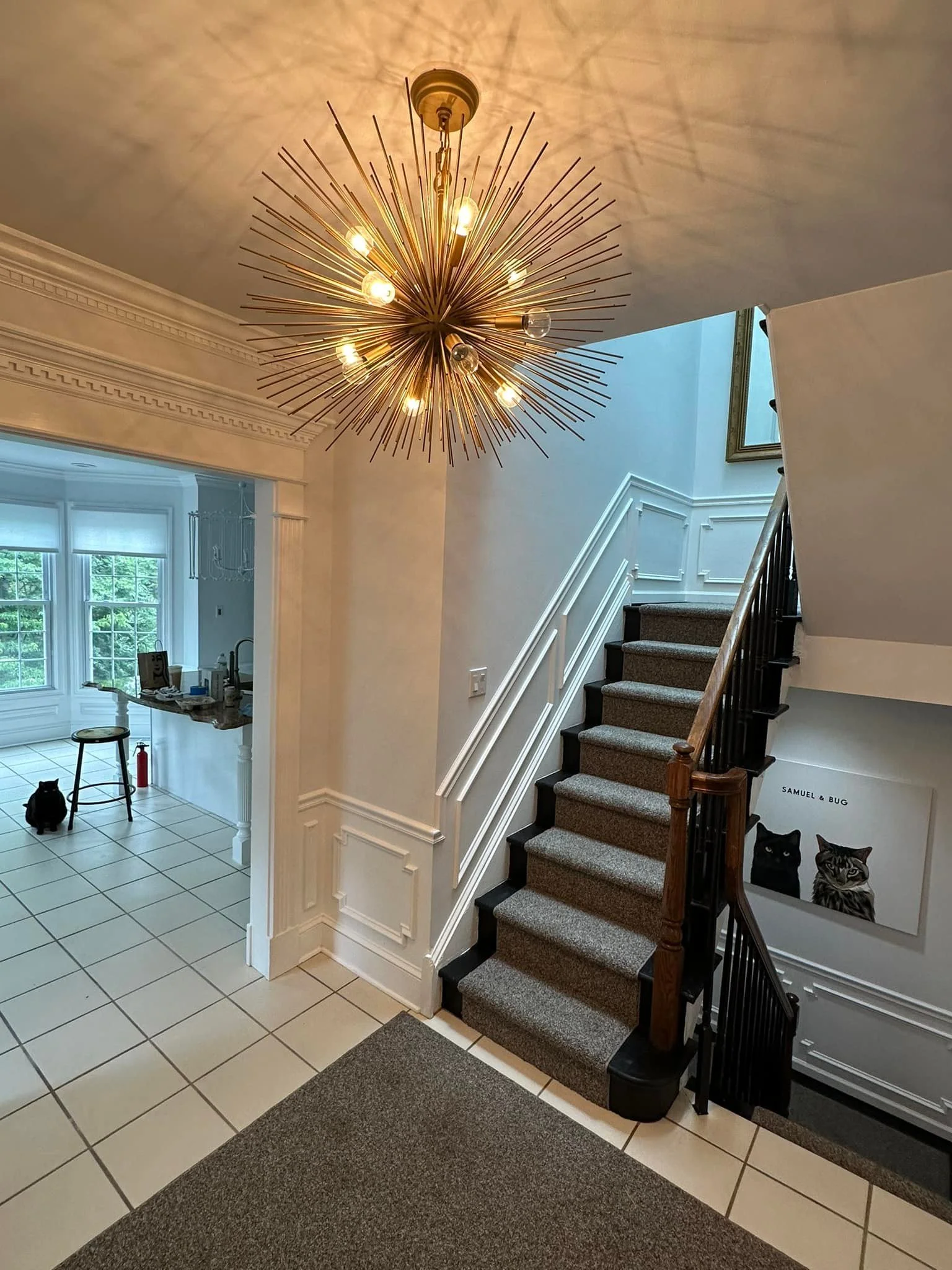 An interior view of a house entryway with a modern starburst chandelier hanging from the ceiling, a staircase with dark carpeted stairs and white railings, a picture of two cats labeled 'Samuel & Bug' on the wall, and a tiled floor leading into a bri