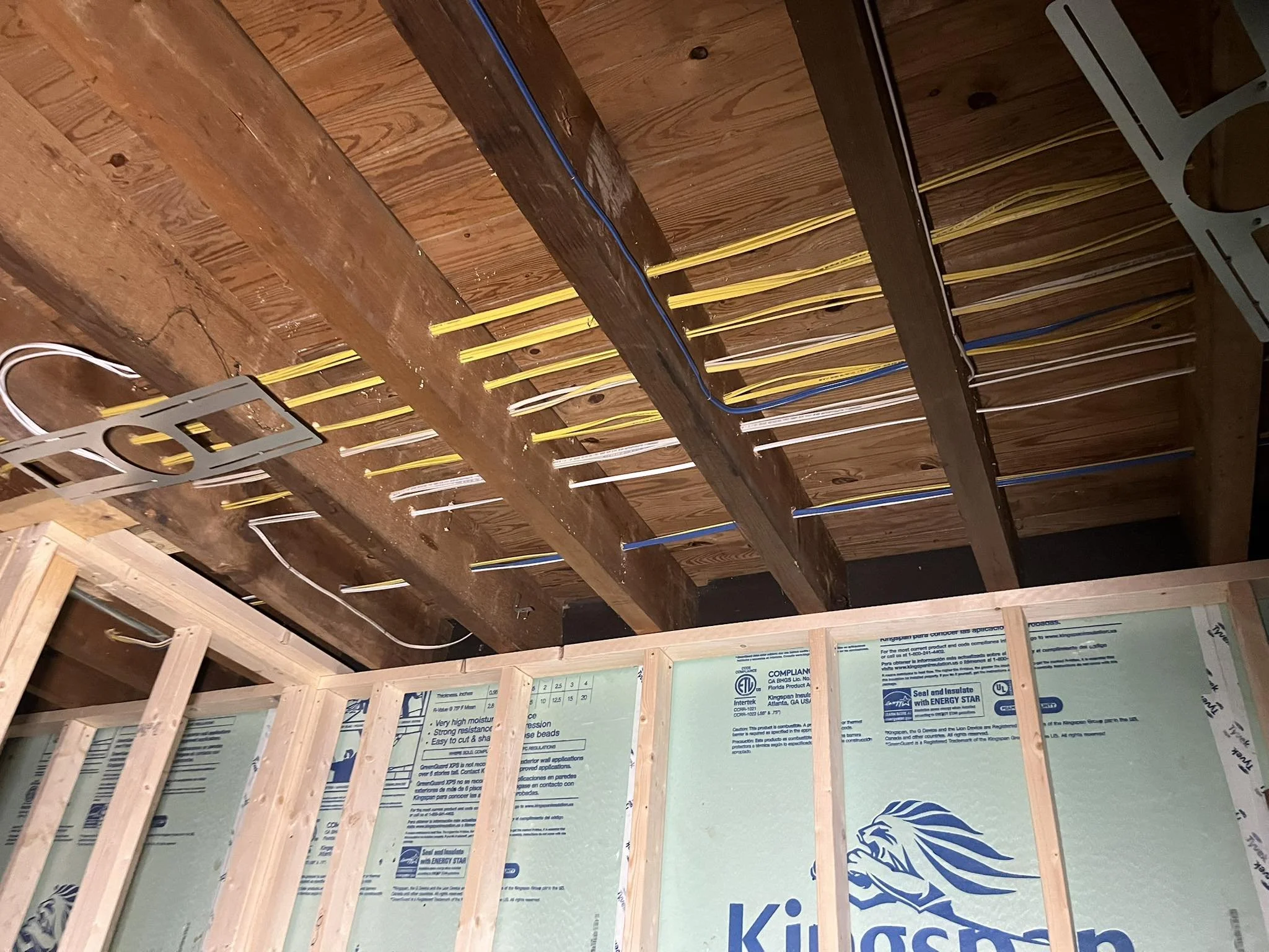 Interior view of an unfinished room showing exposed wooden ceiling beams, electrical wiring, and partial framing with drywall and insulation.