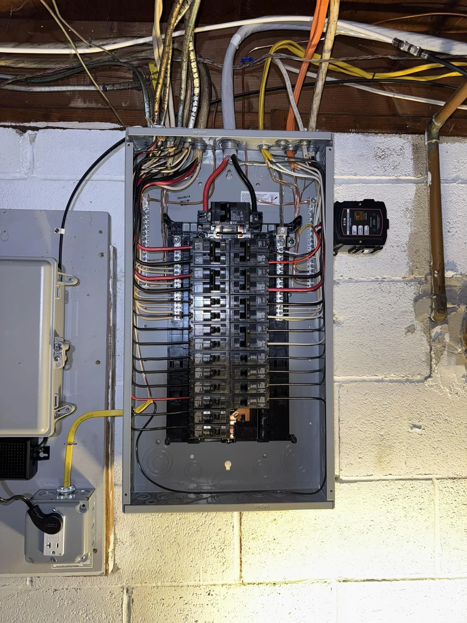 An open electrical breaker box with multiple circuit breakers, wires, and cables connected, mounted on a wall with exposed wiring above.