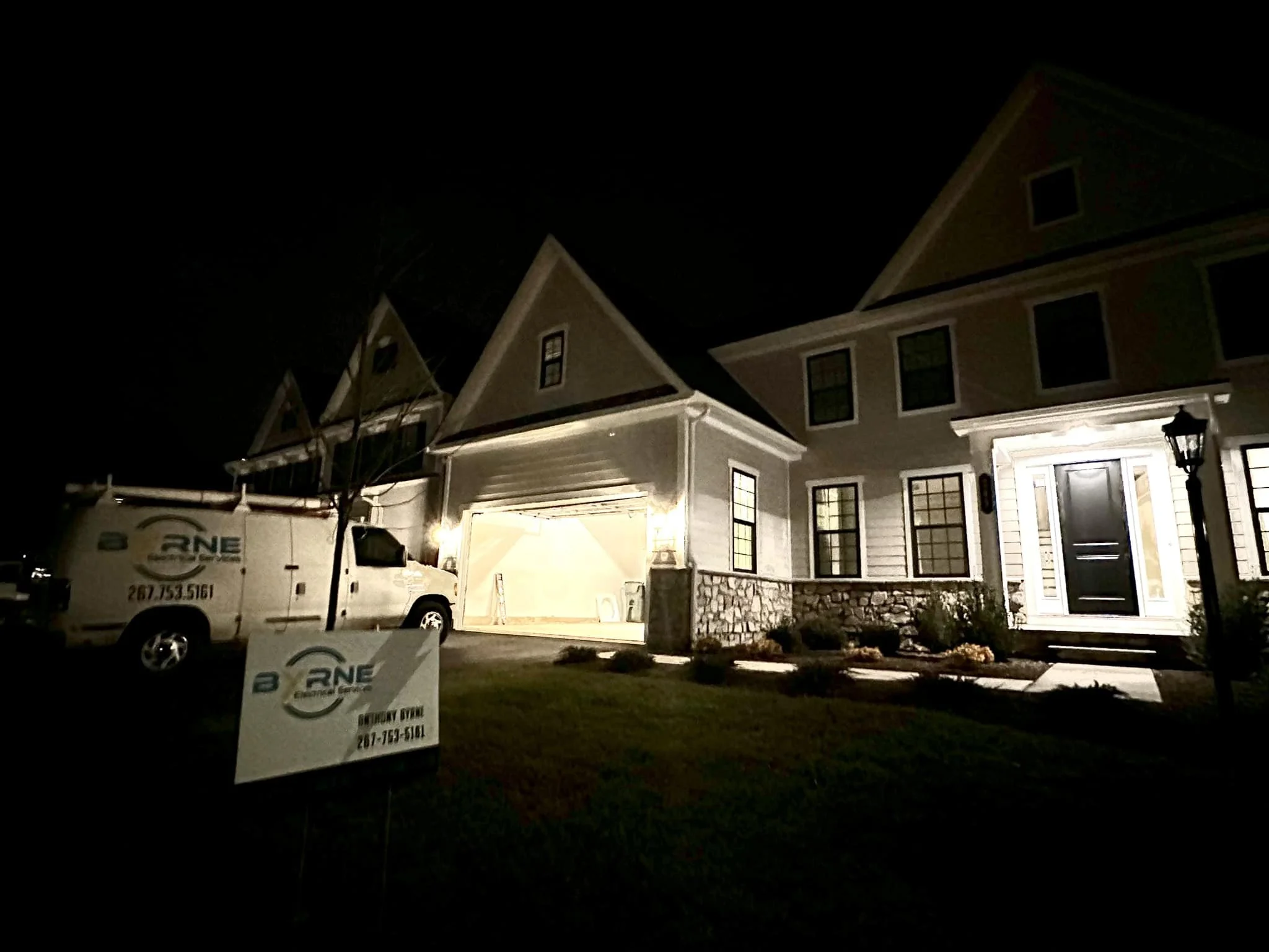 Nighttime photo of a recently constructed house with an open garage, a black front door, lit porch, well-maintained yard, and a sign and truck from a construction company in front.