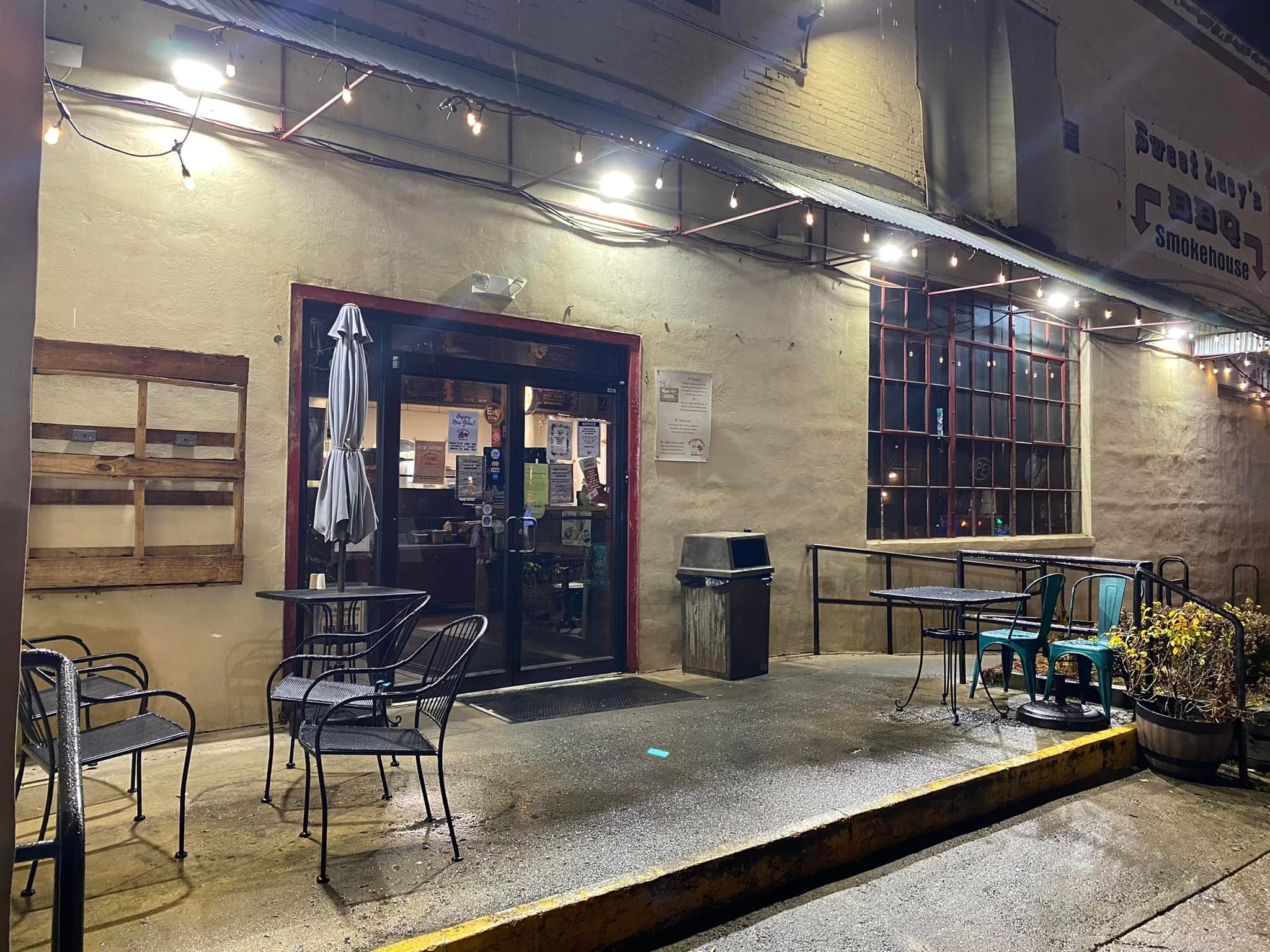 Empty outdoor patio of a restaurant or cafe at night with wet ground, multiple chairs and tables, string lights overhead, a large window with lights inside, a trash can, and a potted plant.