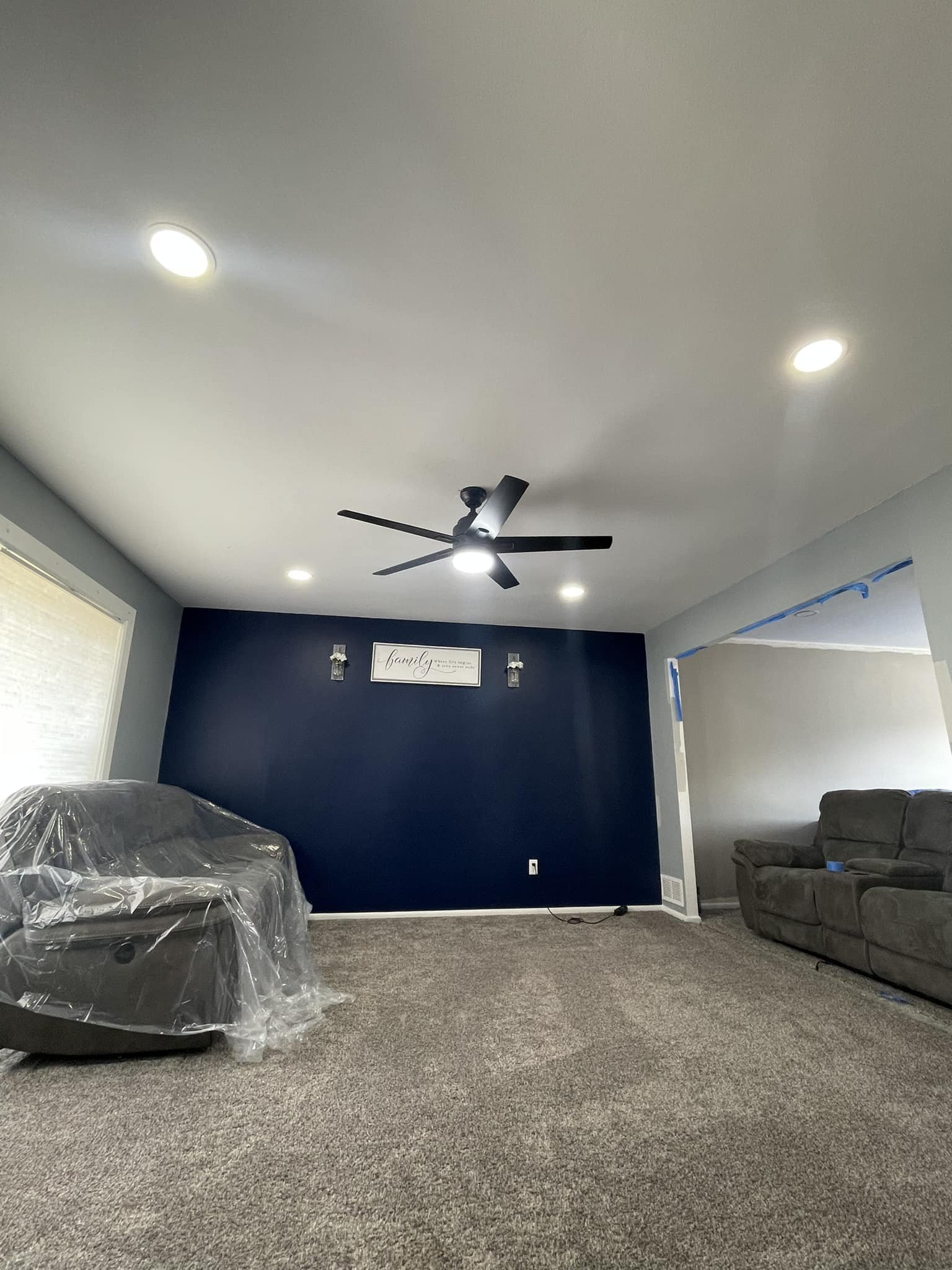 Living room with a dark blue accent wall, beige carpet, a ceiling fan with lights, and two couches, one covered in plastic. There is a wall sign that says "family" and small picture frames on the wall.