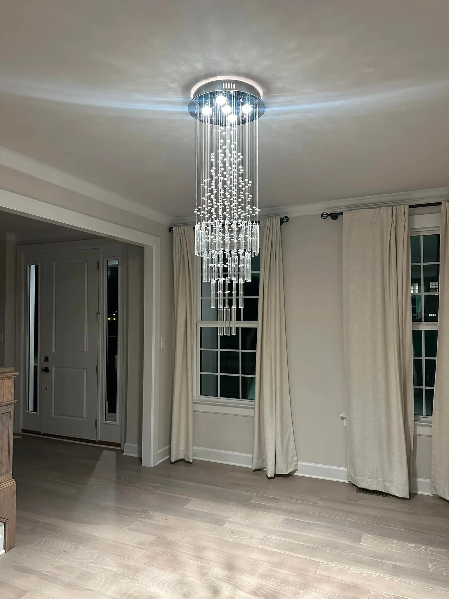 Modern chandelier hanging from the ceiling above a room with light-colored hardwood floors, white walls, and cream curtains. Night outside the window.