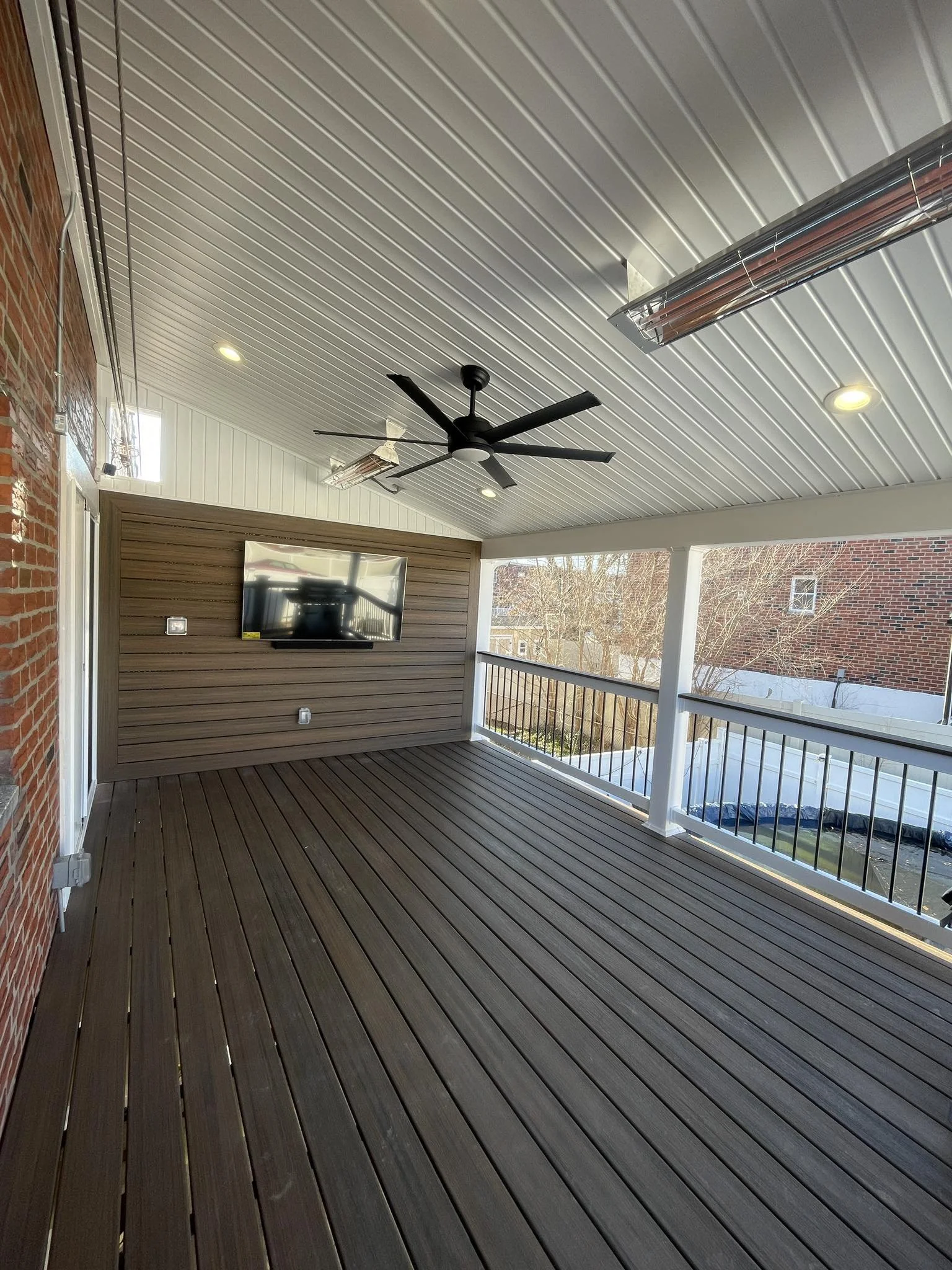 Spacious balcony with wooden floor planks, a wall-mounted flat-screen TV, ceiling fan, modern ceiling heater, and railing overlooking neighboring houses with clear sky.