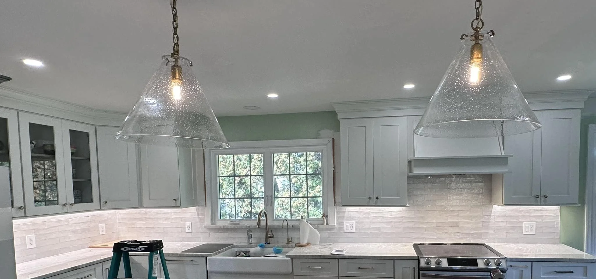 Kitchen with light green cabinets, a white subway tile backsplash, a window above the sink, and two glass pendant lights hanging from the ceiling.