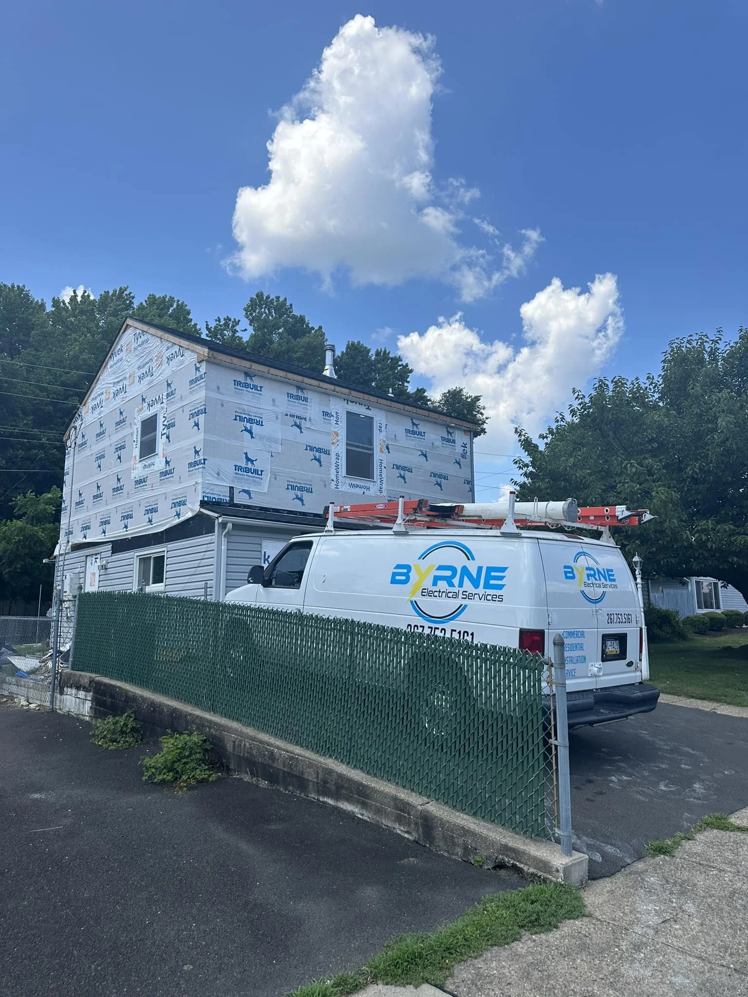 House under construction with siding partially installed, parked with a BYRNE Electrical Services van with ladder on top, in a residential neighborhood with trees and a blue sky with clouds.