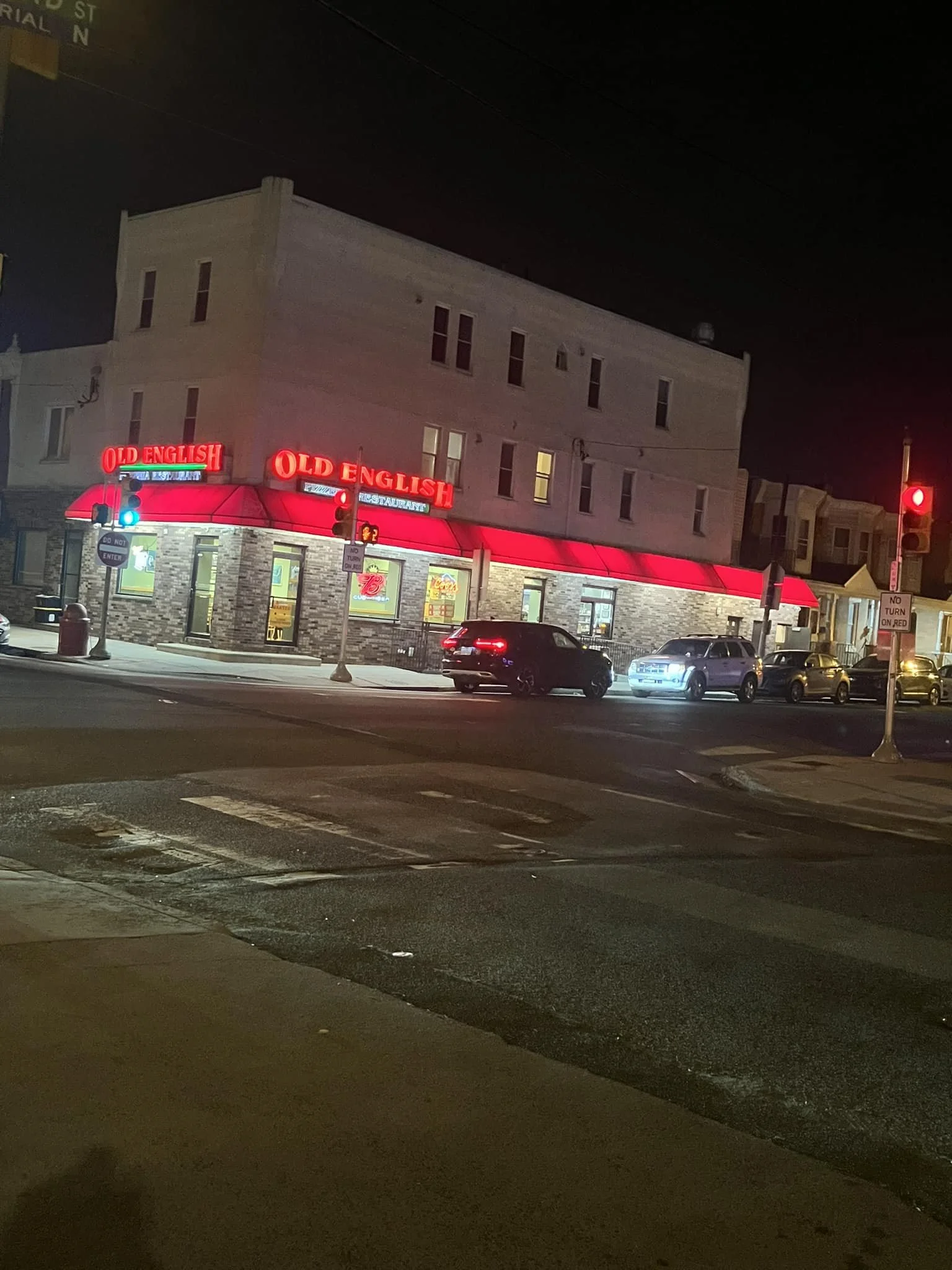 Nighttime street scene with a restaurant named 'Old English' featuring a red awning and illuminated neon signs. Several cars are parked and driving past, with traffic lights visible.
