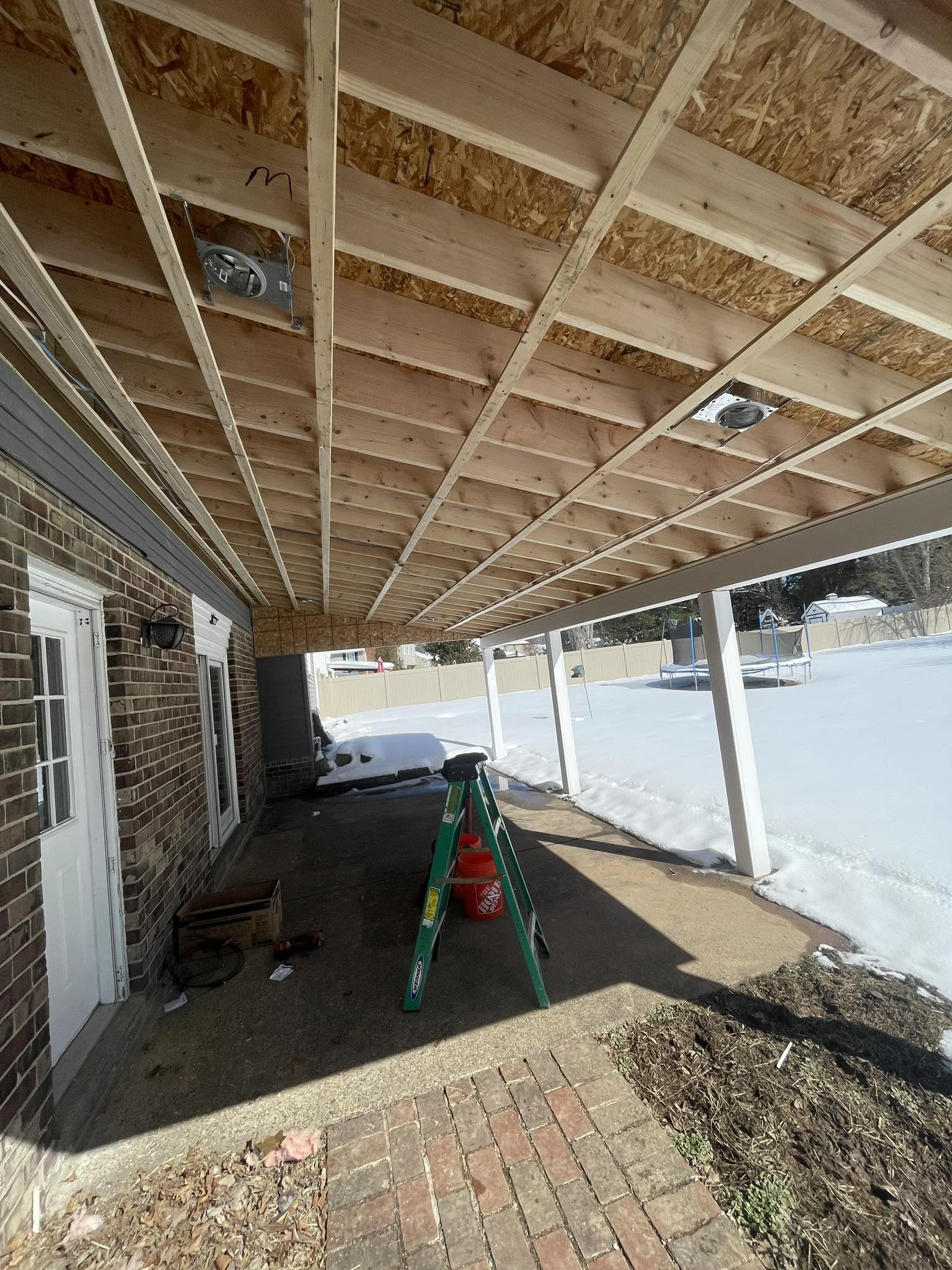 View of a house porch under construction with exposed wooden beams and a roof, with snow-covered yard and trampoline in the background.