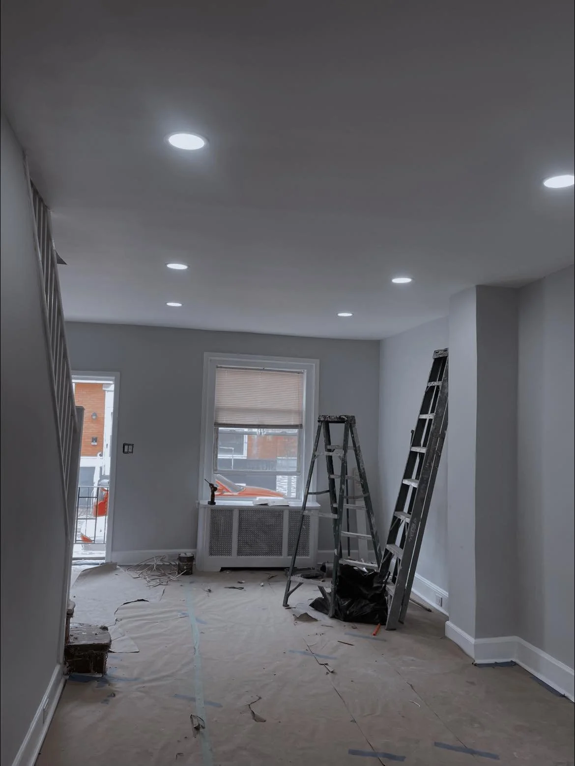 Living room under renovation with ladders, plastic covering on the floor, and construction tools near the window.