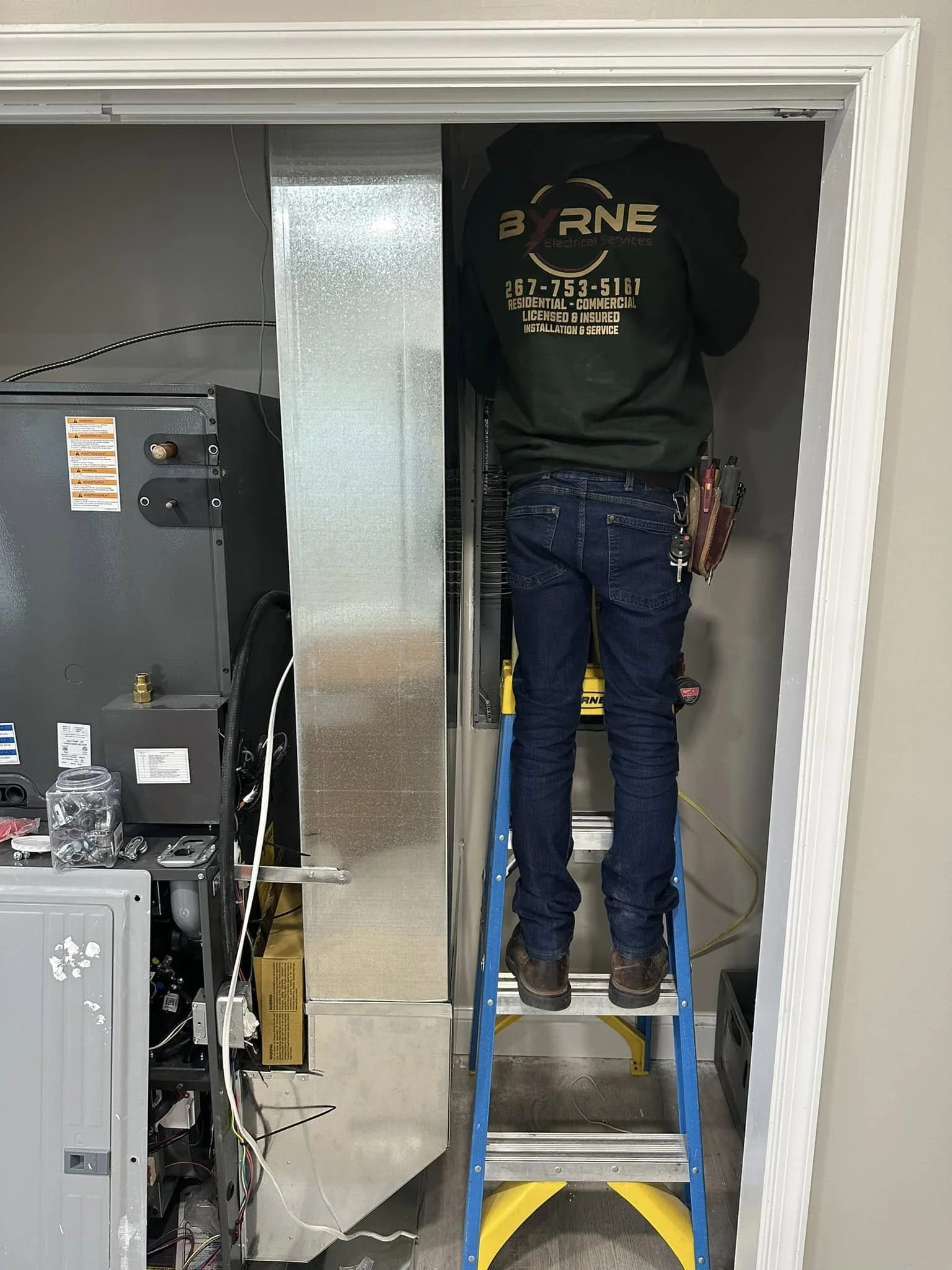 Technician standing on a small stepladder working inside an HVAC or electrical service closet, wearing a jacket with company logo and phone number.