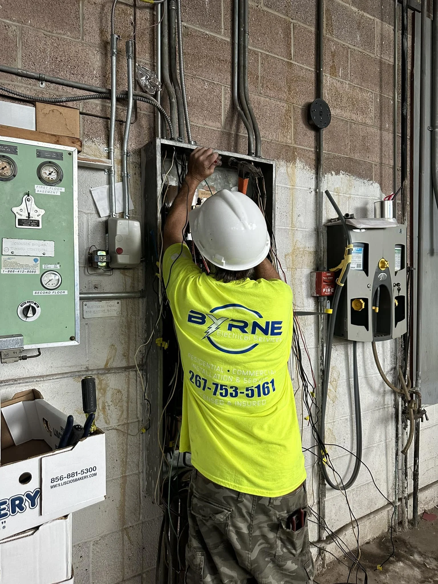 An electrician working on wiring inside an electrical panel on a wall, wearing a white hard hat and a neon yellow shirt with The Byron Electrical Services logo.