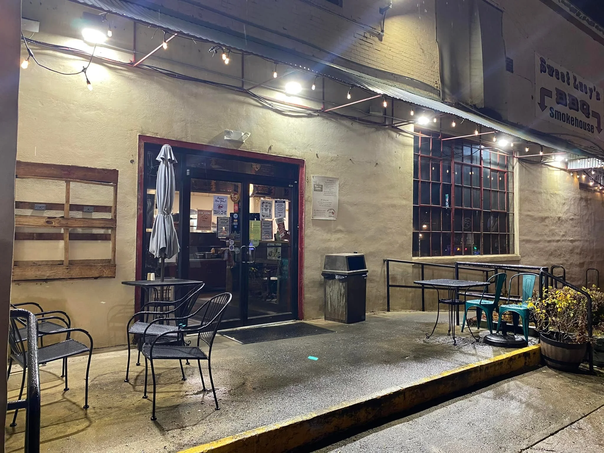 Exterior of a cozy cafe or restaurant at night with outdoor seating, string lights overhead, and a large window showing the inside. There are a few tables and chairs outside, some with umbrellas, and a trash can near the entrance.
