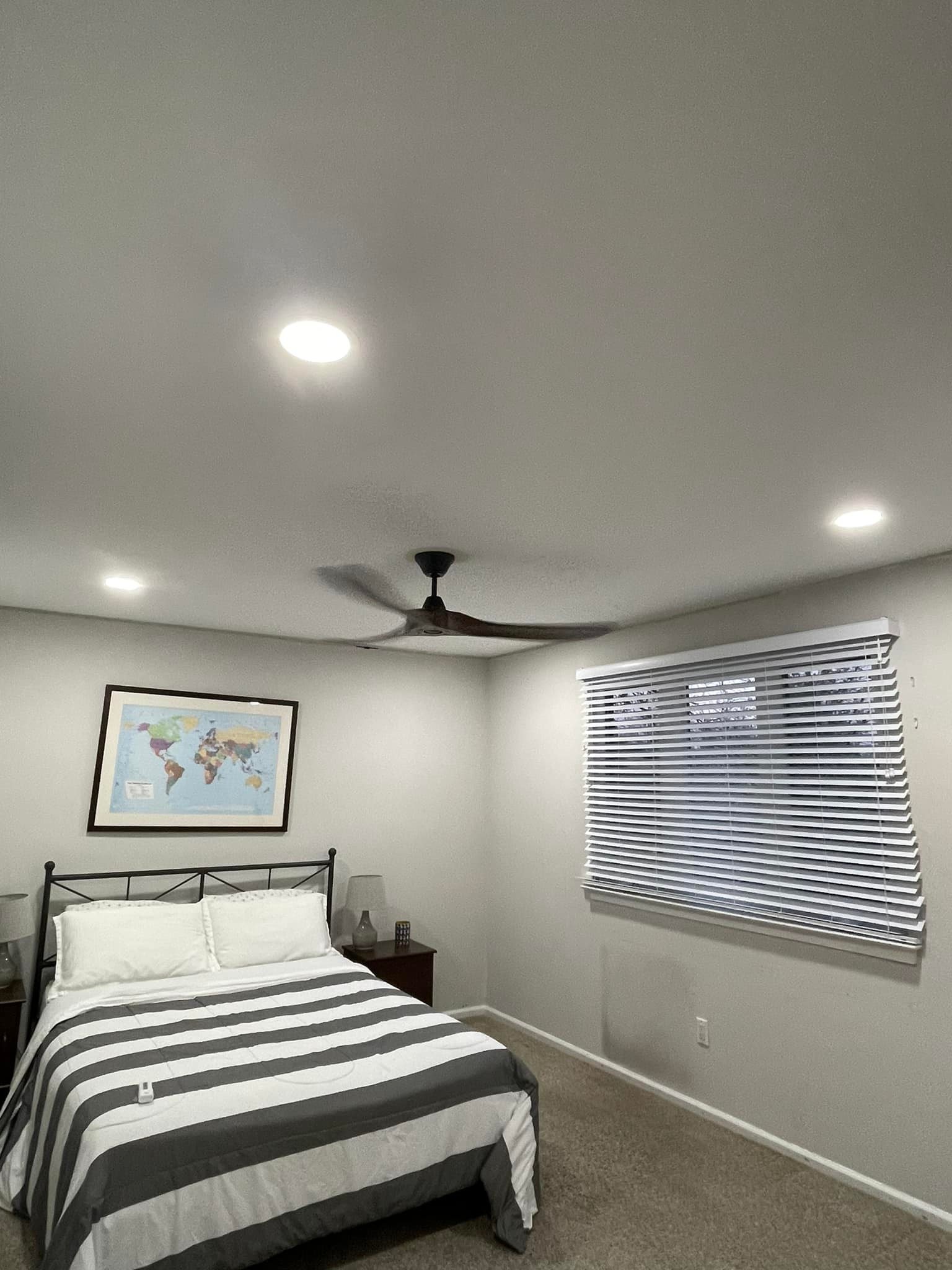 A bedroom with a bed with striped gray and white bedding, two bedside tables with lamps, a framed world map on the wall, a window with horizontal blinds, a ceiling fan, and recessed lighting.