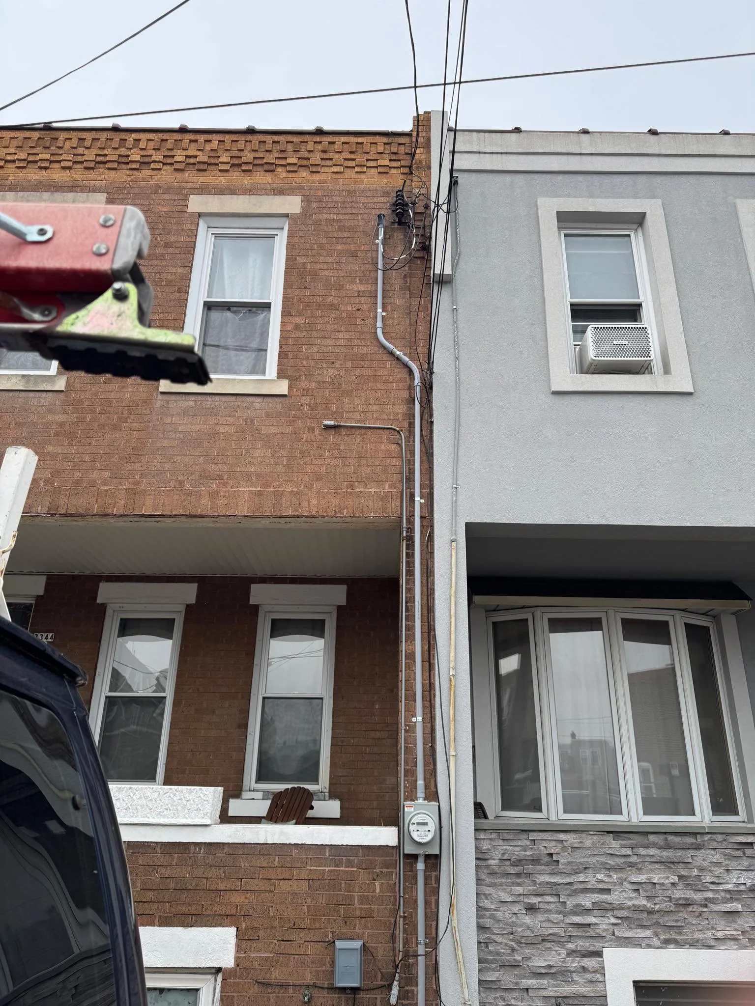 View of two adjacent urban residential buildings with different exterior finishes, windows, and visible utility meters and wires on a cloudy day.