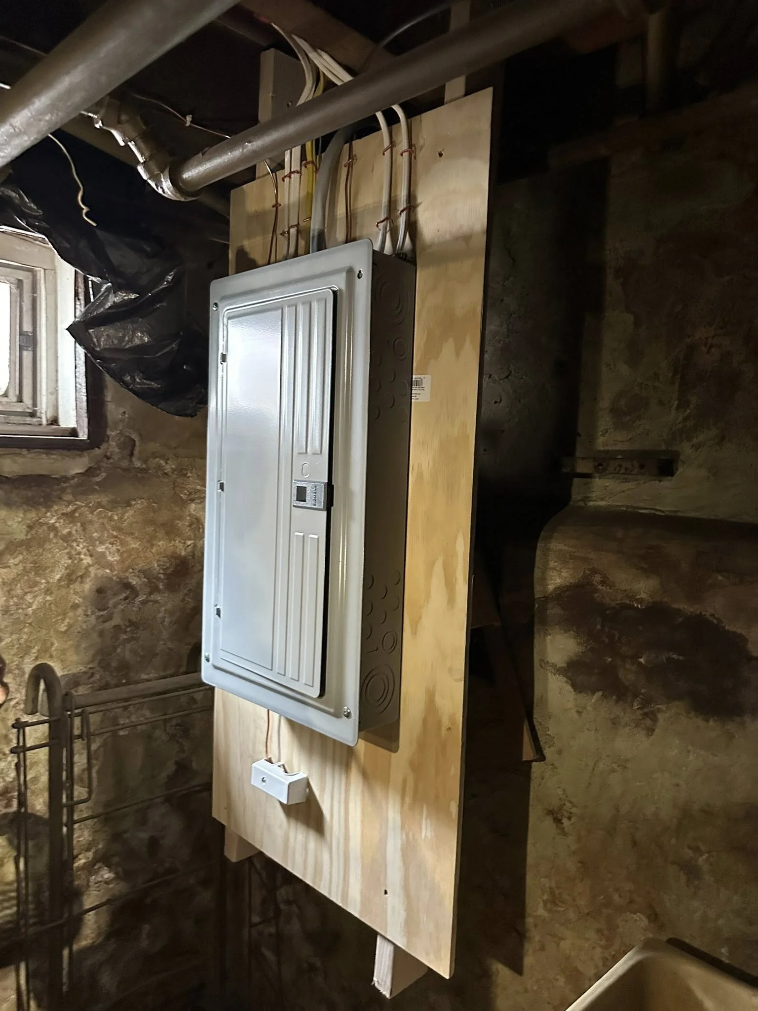 A metal electrical panel mounted on a wooden board in a basement or utility room. The panel has a wire conduit leading into it, and a small box or door on the front. The room has stone walls and a small window.