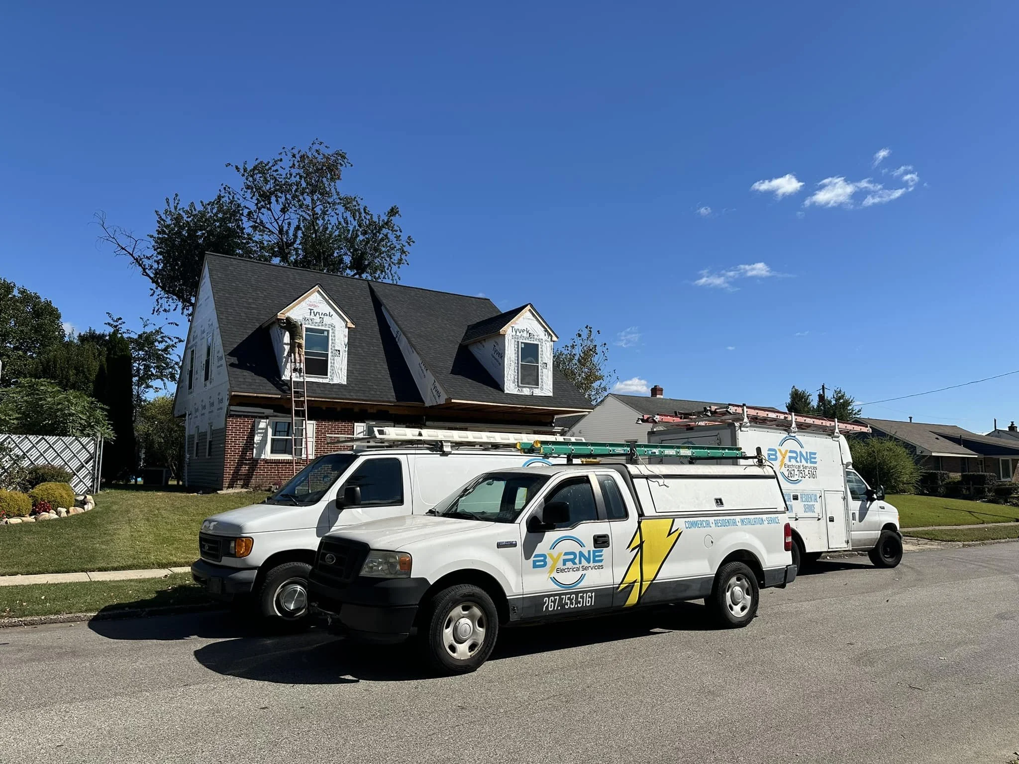 Construction workers installing a roof on a house, with two company vans parked in front on a sunny day.