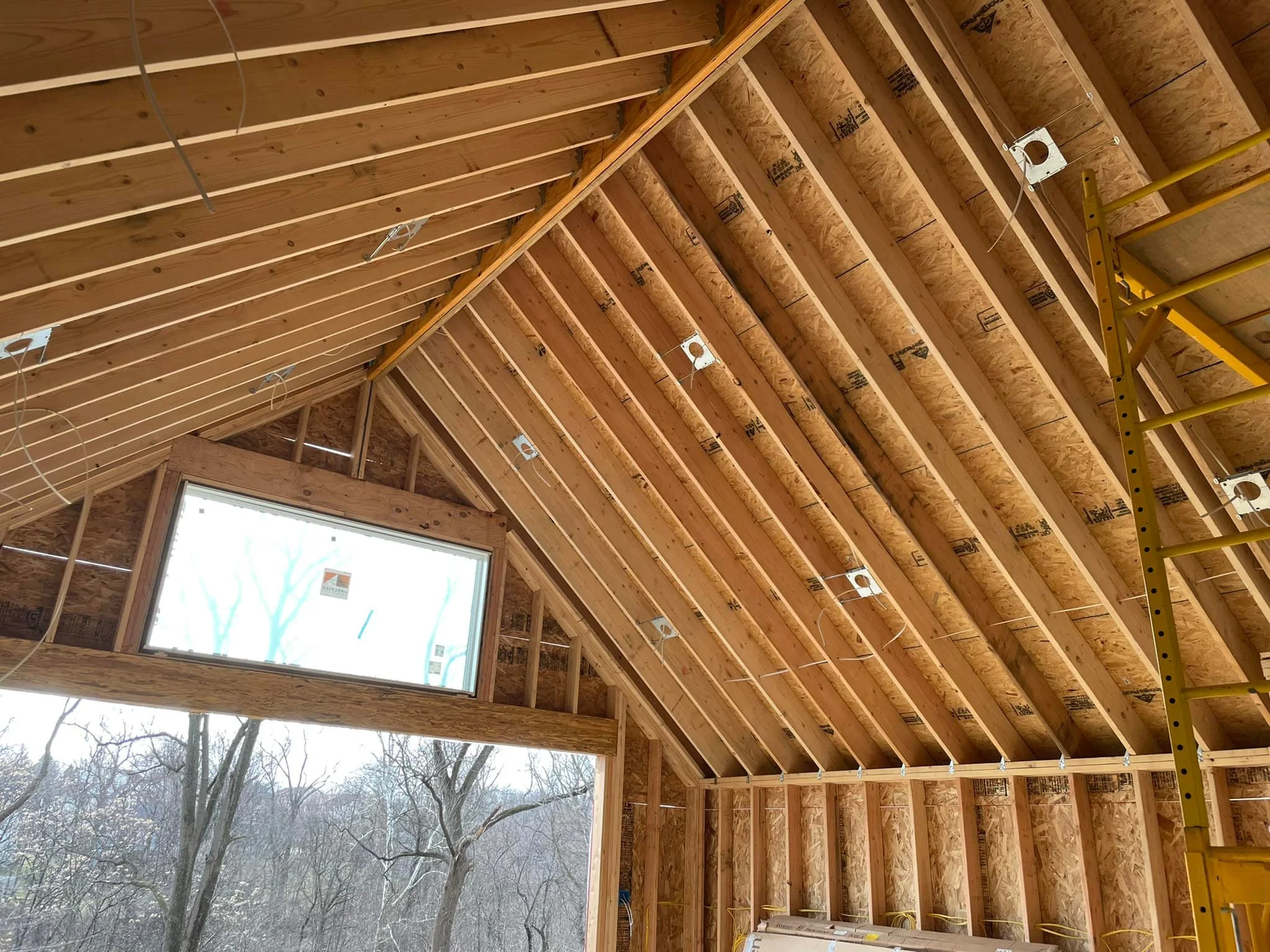 Interior of a building under construction with exposed wooden framing, electrical wiring, and a large window overlooking trees outside.