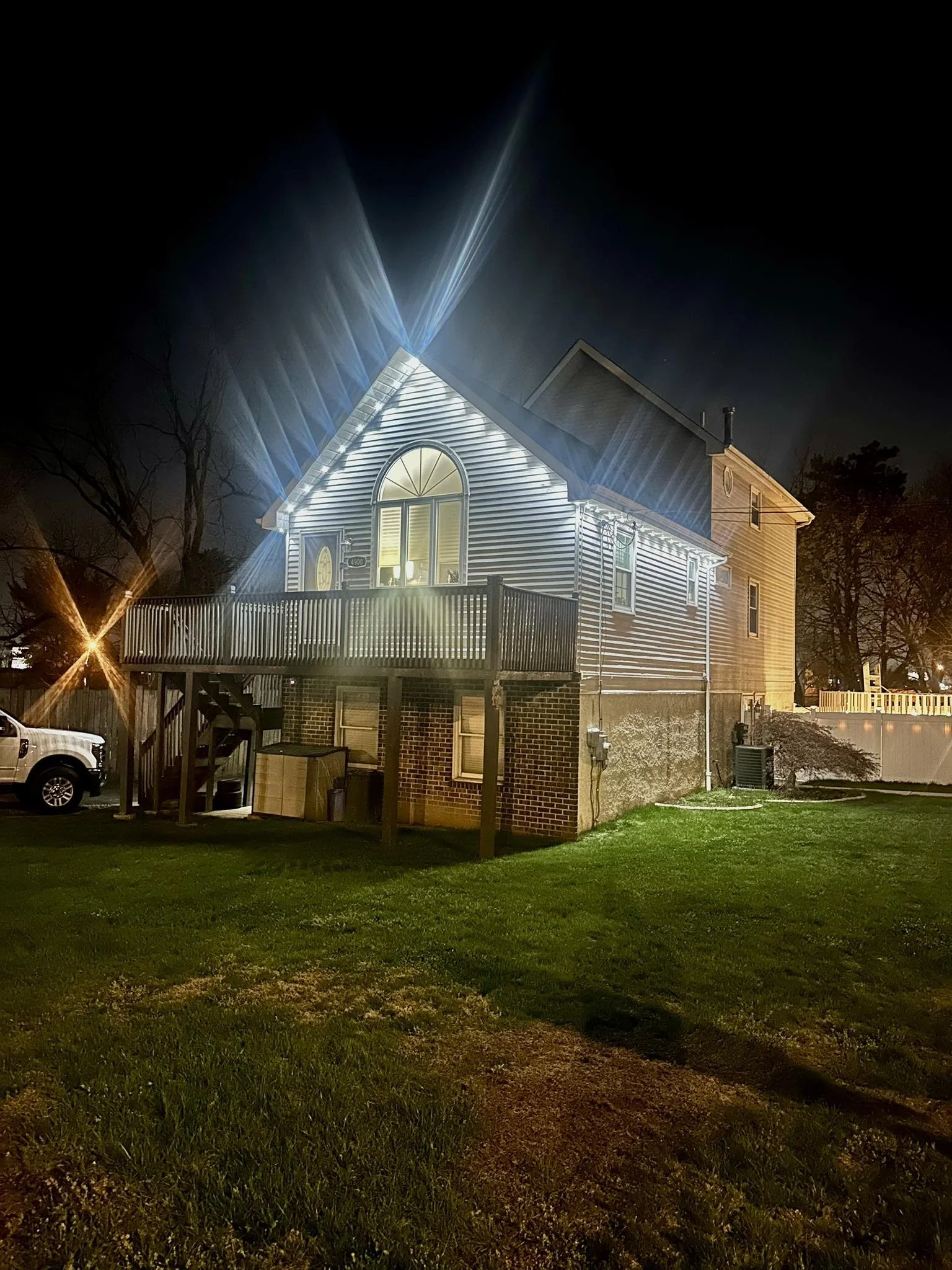 Nighttime photo of a two-story house with a lit-up upper floor, a deck with stairs, and a lawn in the foreground. Bright lights create starburst effects, illuminating the house and surroundings.