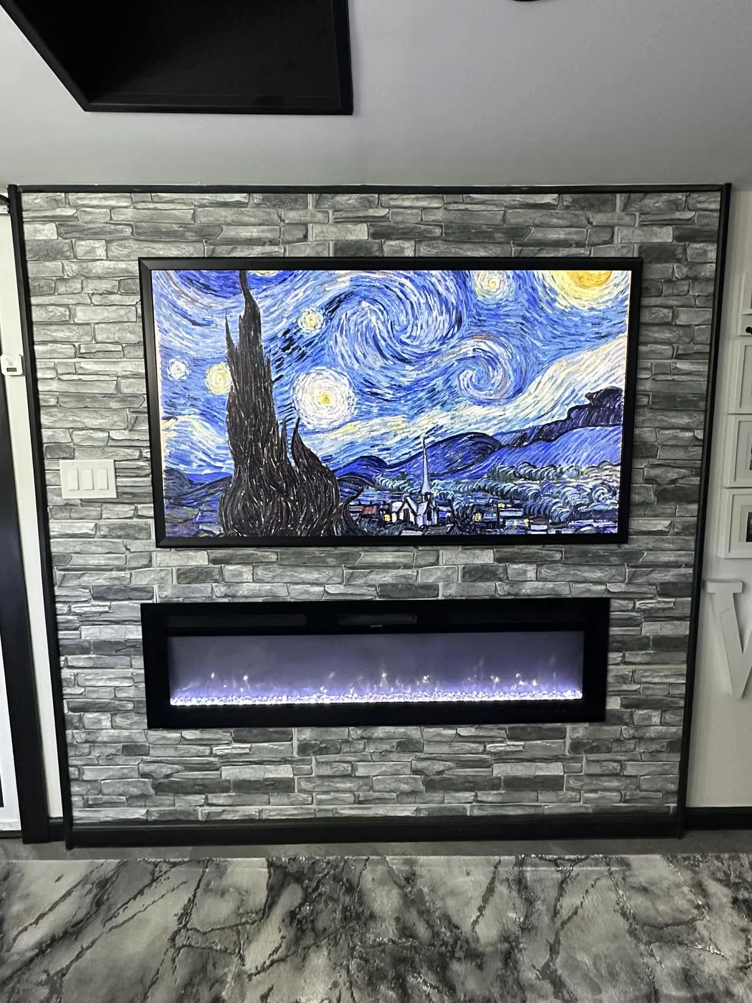 Photo of a room with a large TV displaying Vincent van Gogh's Starry Night painting above a modern electric fireplace with a long rectangular shape and visible flames, all set against a brick wall.