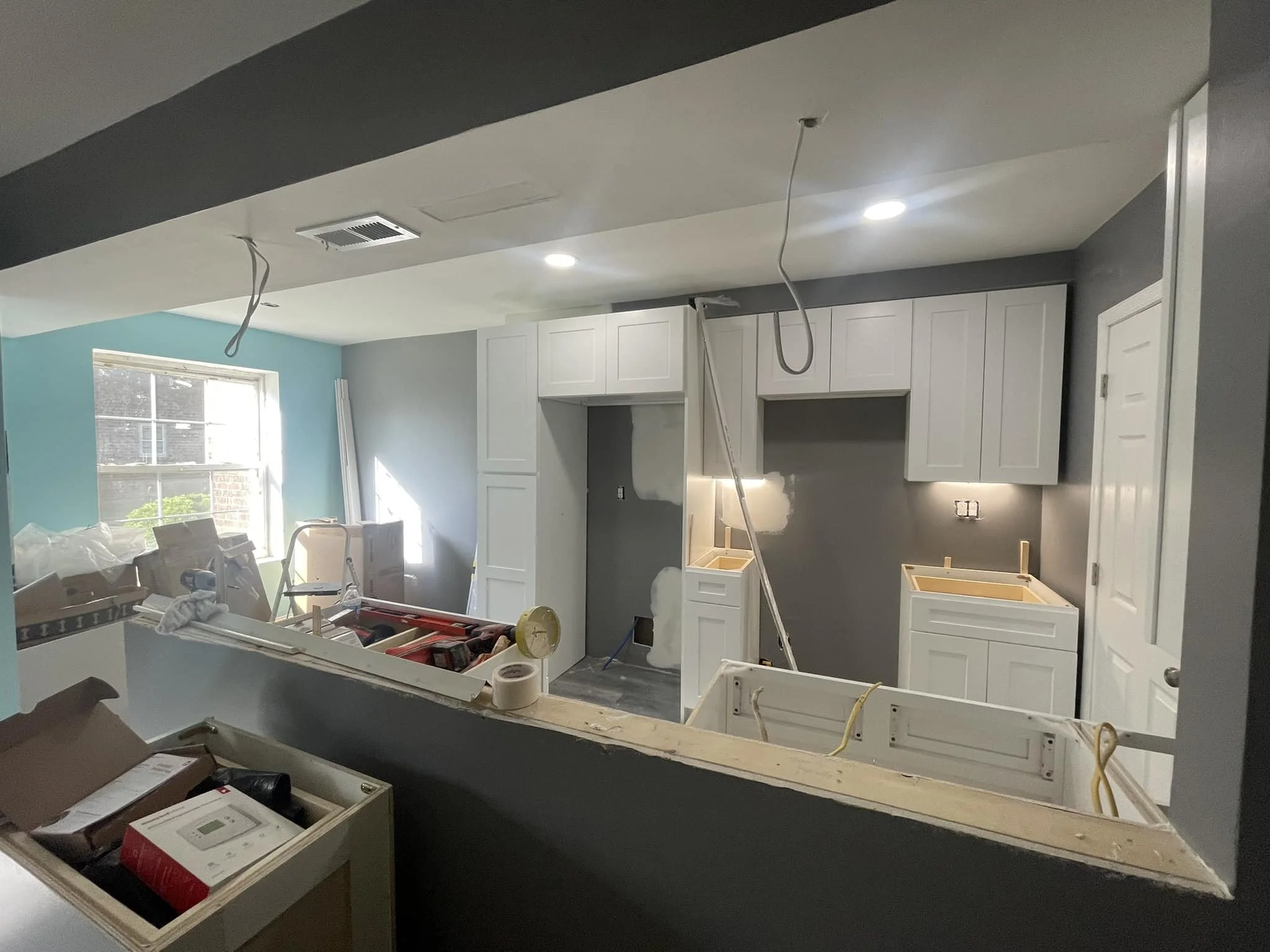 Kitchen under renovation with white cabinets, gray walls, and construction tools and materials scattered around.